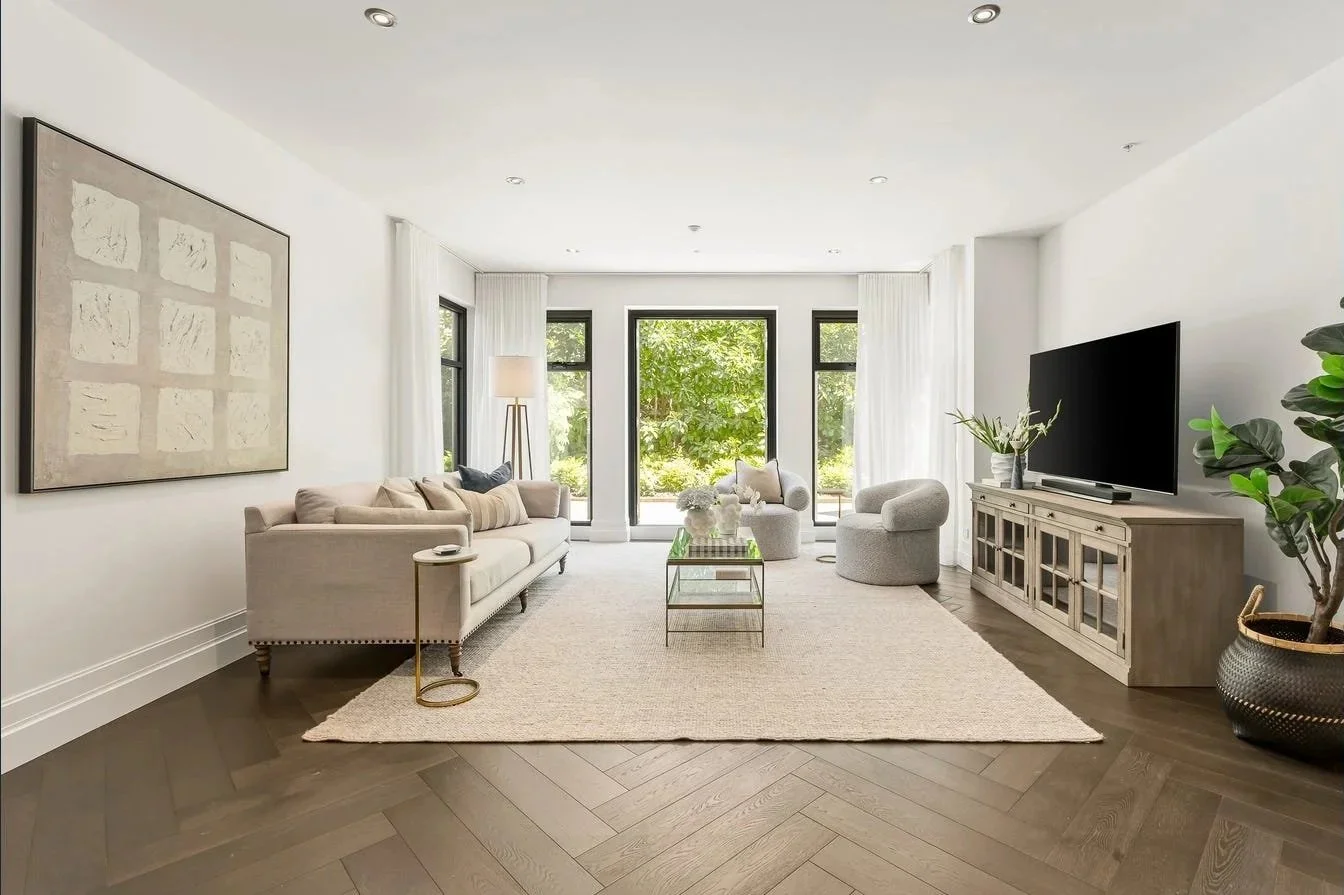 Bright living room with white walls, large windows, and minimalist furniture including a sofa, armchair, coffee table, and TV cabinet. Decor includes artwork, potted plant, and a beige rug.