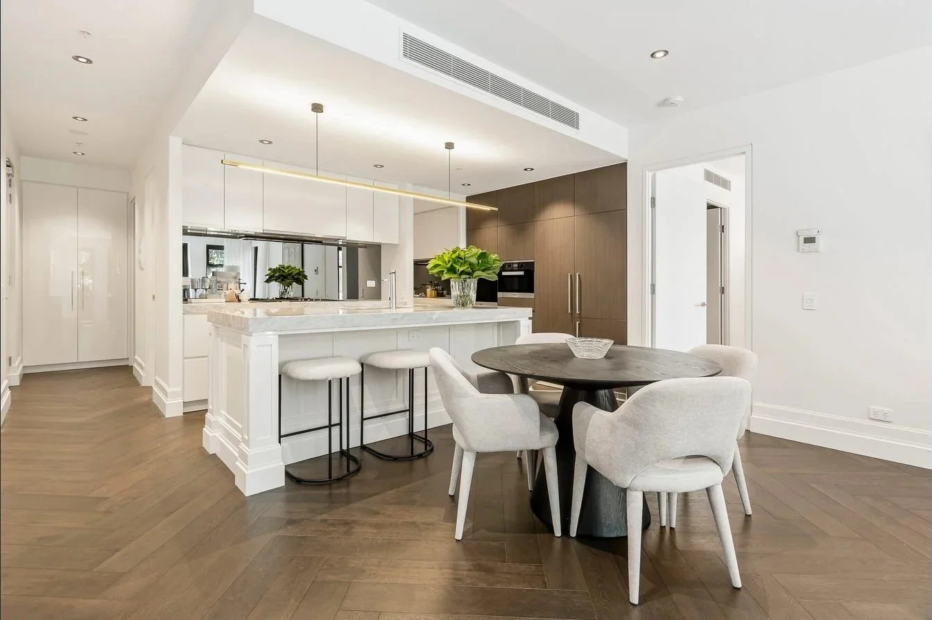 Modern open-concept kitchen and dining area with white walls, wooden floors, a round dining table surrounded by four light-colored chairs, an island with barstools, and contemporary cabinetry.