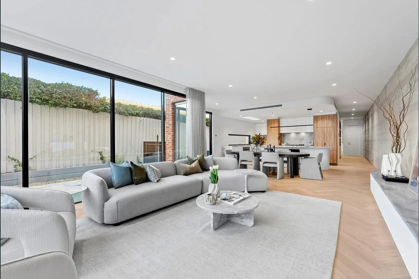 Open-concept living room and kitchen with large glass windows, light-colored furniture, wooden flooring, and modern decor.