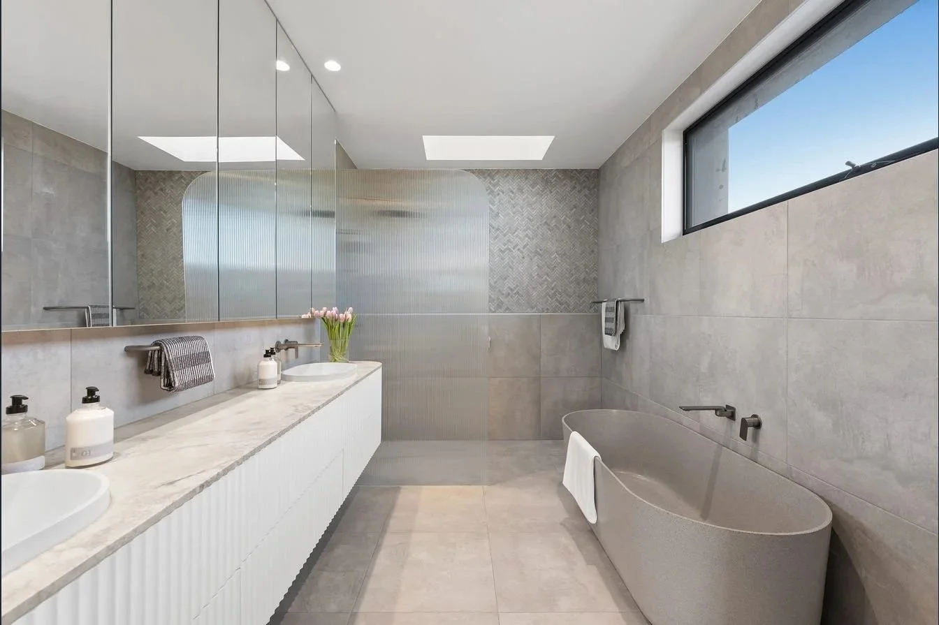 Modern bathroom with a neutral color palette, large wall mirror, double vanity with toiletries, a vase of pink tulips, a soaking bathtub with a towel, and a window letting in natural light.