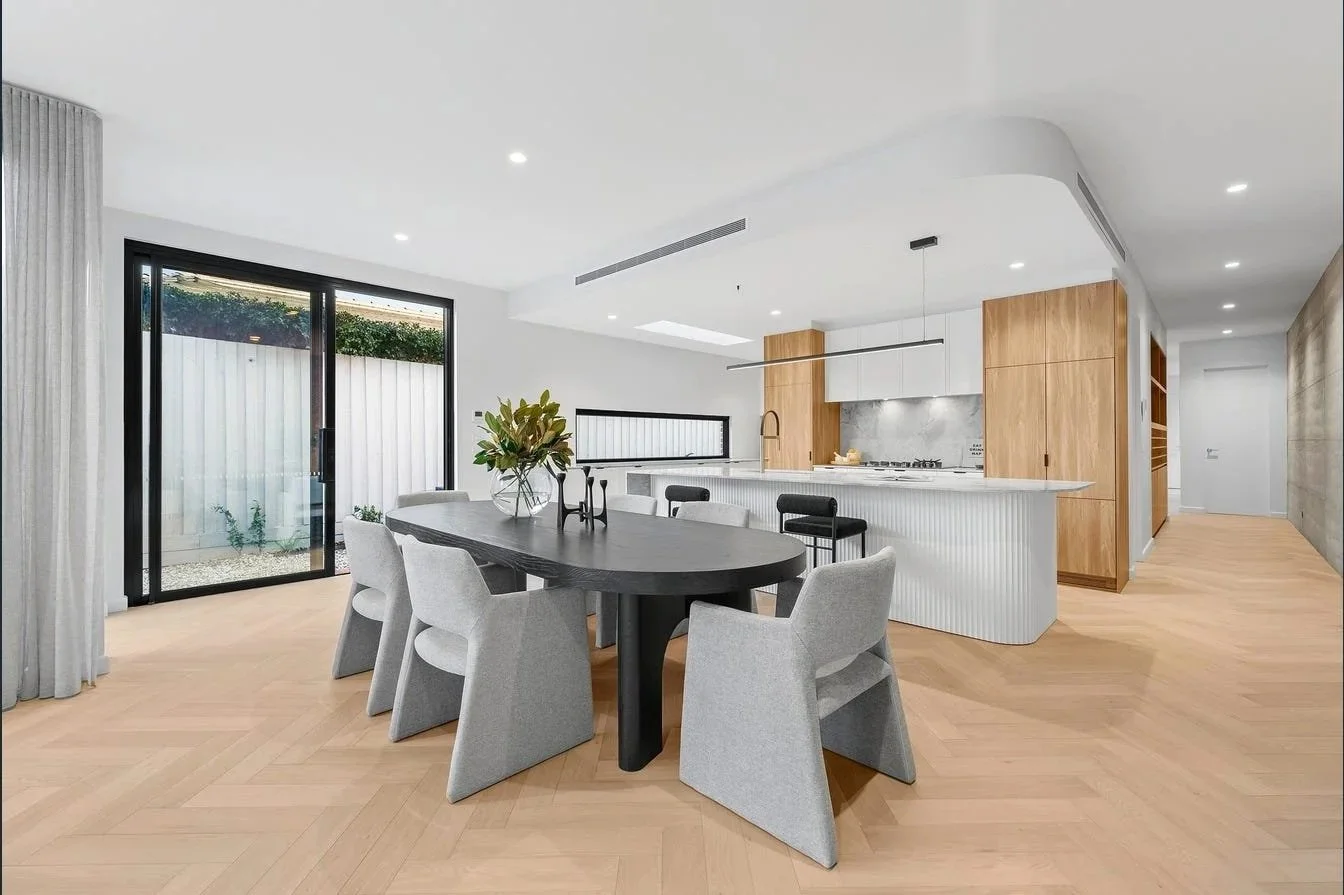 Modern open-concept kitchen and dining area with a large black dining table, gray chairs, and a kitchen with white cabinets, wooden accents, and a marble backsplash. Large sliding glass door with curtains leads outside.