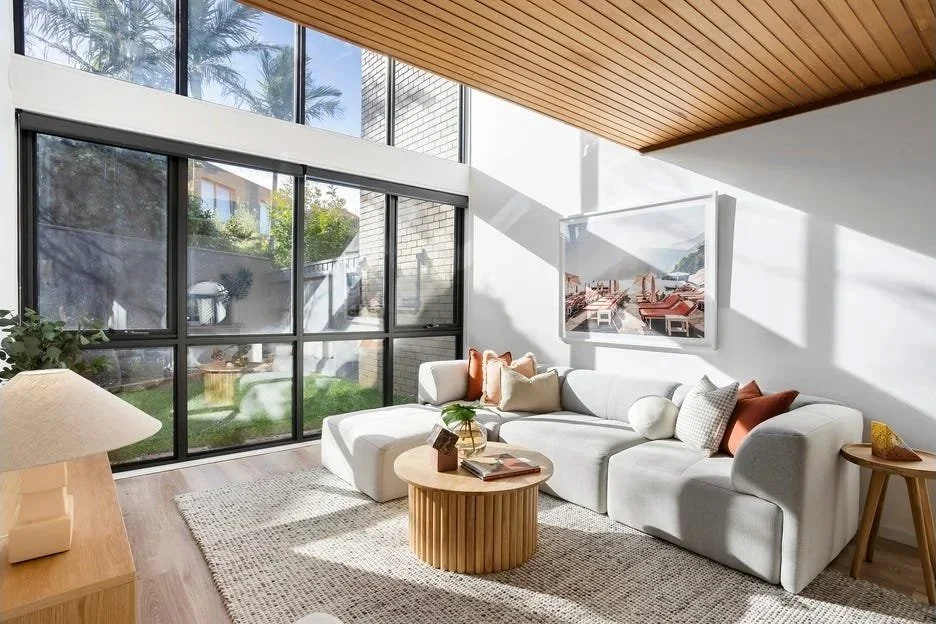 Bright living room with large floor-to-ceiling windows, white sectional sofa with pillows, round wooden coffee table, artwork on the wall, and wooden ceiling panel.