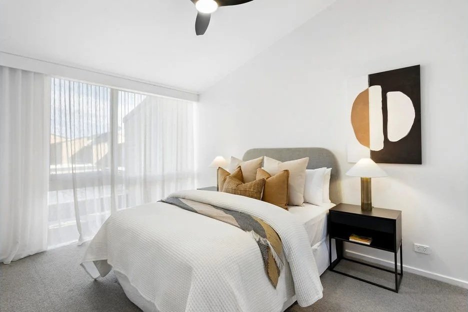 Bright bedroom with a large bed, white bedding, multiple pillows, and a bedside table with a lamp. Large windows with sheer curtains, and modern minimalist art on the wall.