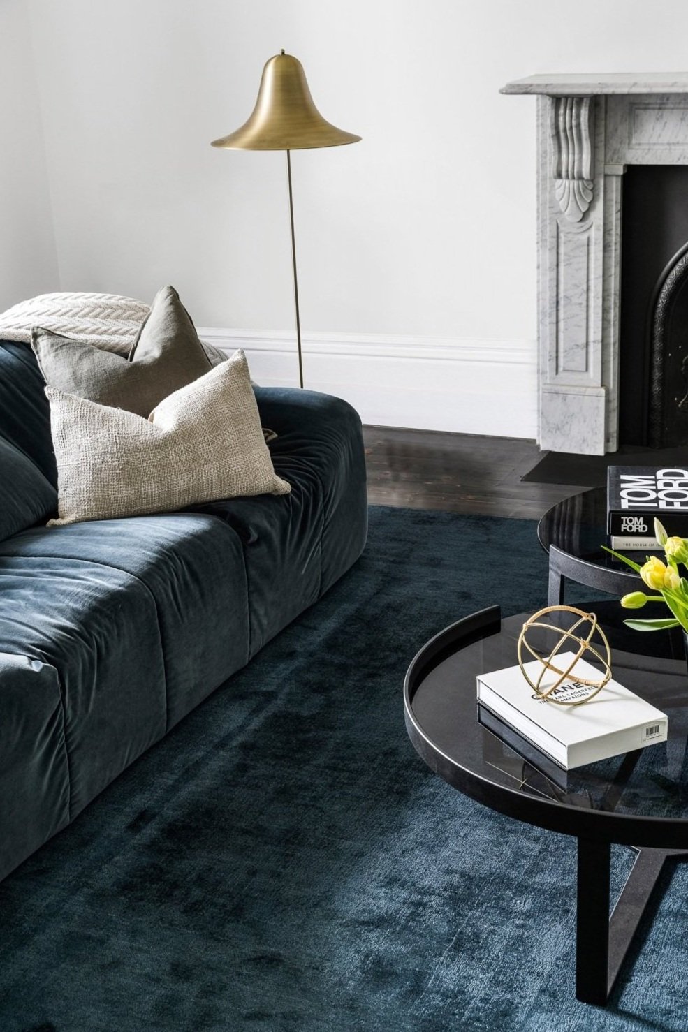 Living room corner with a black velvet sofa, beige throw pillows, a gold floor lamp, a white marble fireplace, a round black coffee table with books and decorative objects, and a dark blue rug.