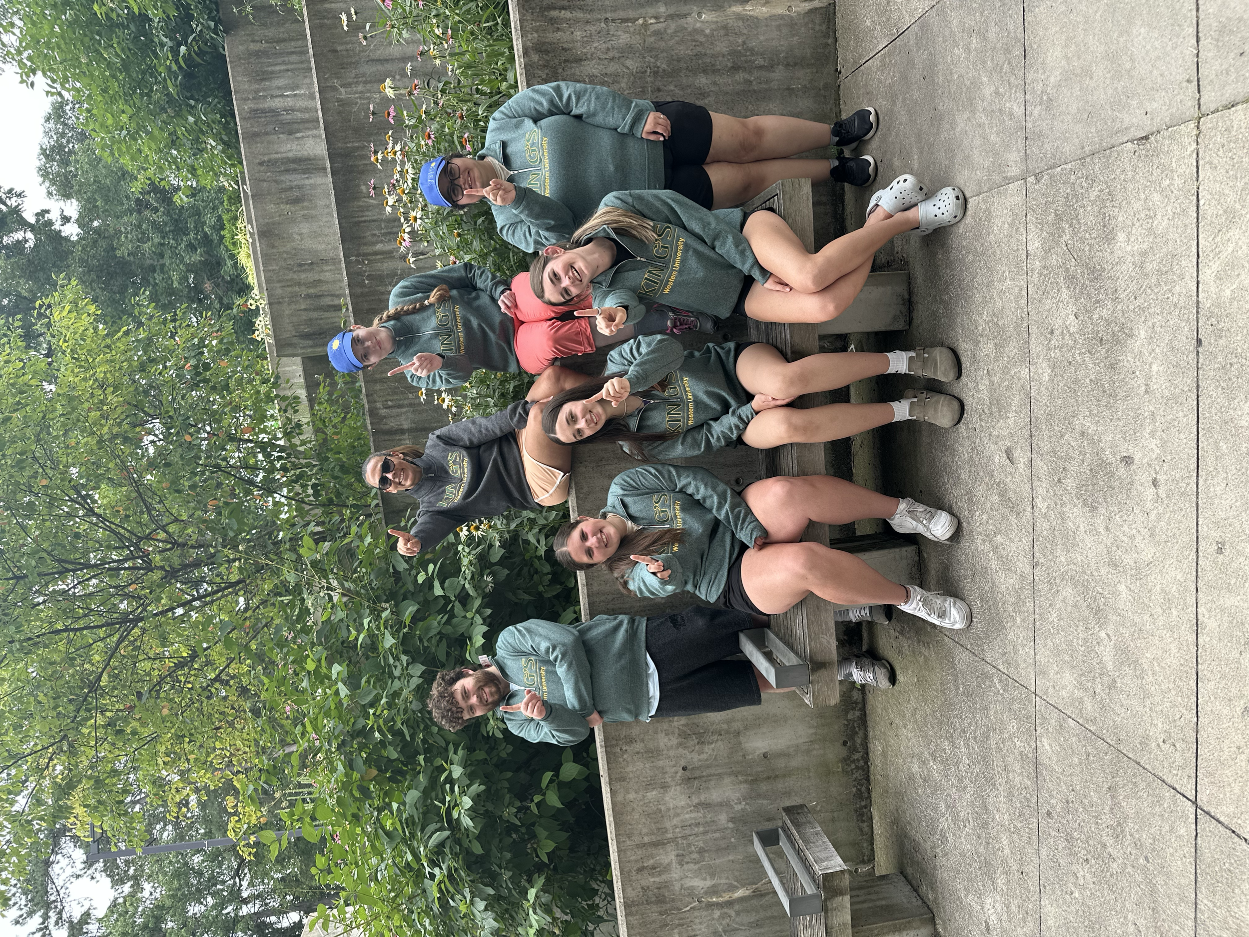 Group of six young women and two young men from Your Neighbourhood Camp some wearing King's University labled sweat shirts , sitting and standing holding up one finger signalling  number 1. The background has trees, greenery, and a concrete block.