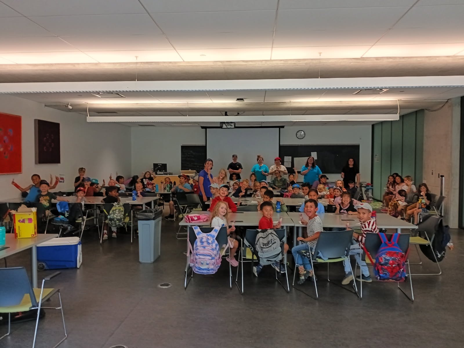 Classroom at Your Neighbourhood Camp filled with children sitting at tables, some waving or smiling, with teachers standing at the back of the room.