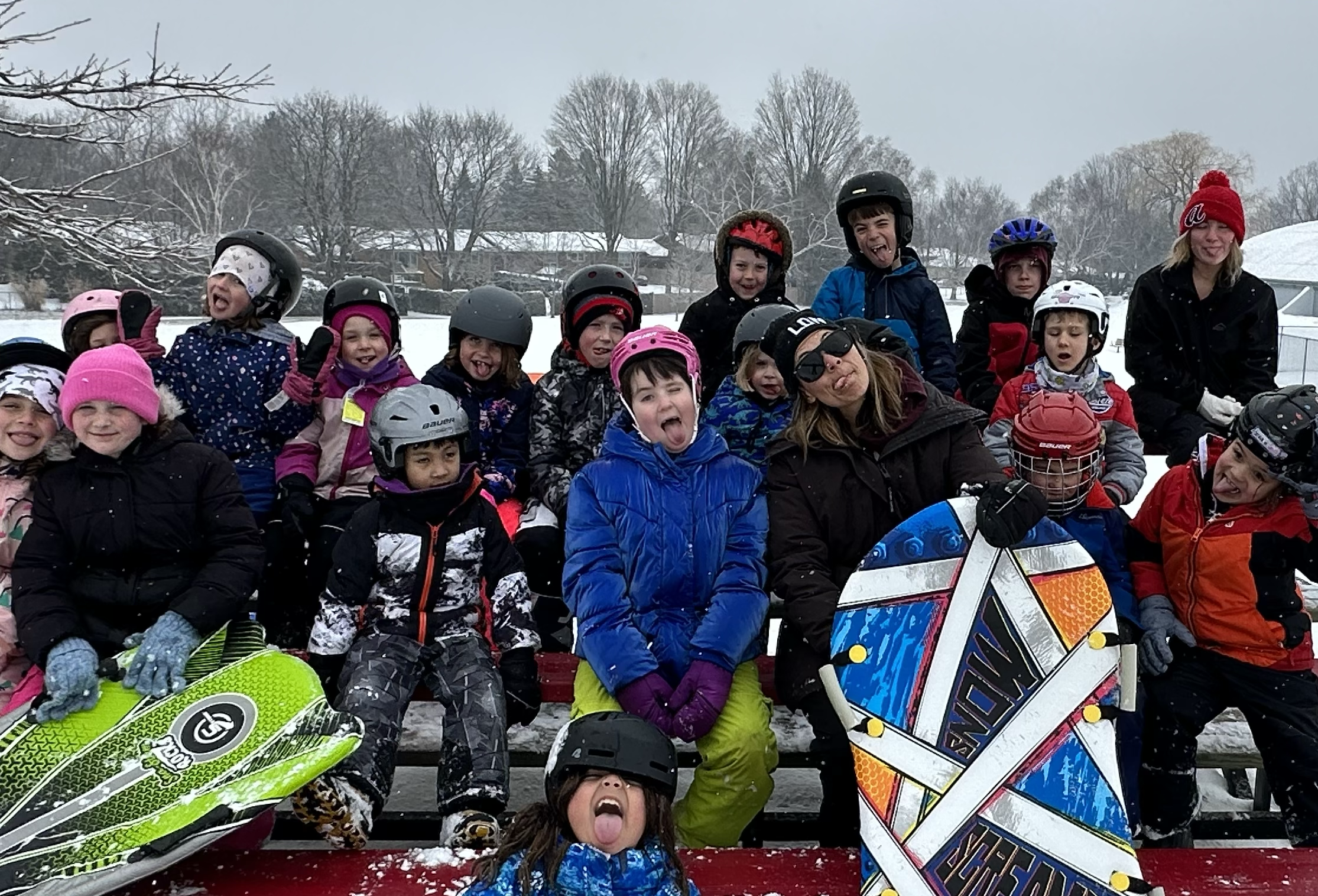 Group of children and staff members from Your Neighbourhood Campin winter clothing and helmets posing with snowboards on a snowy outdoor background.