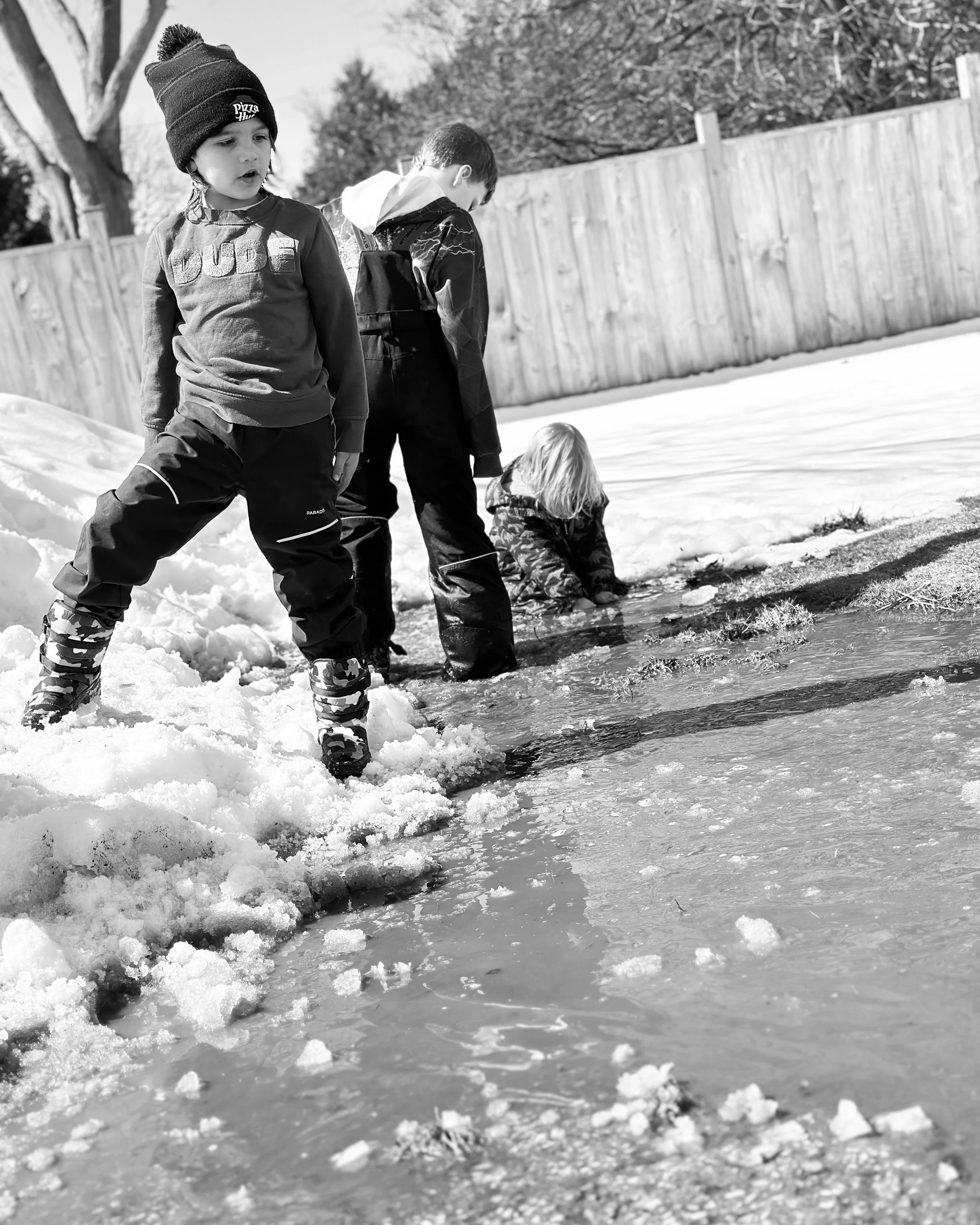 Three children at Your Neighbourhood Camp playing at the edge of a frozen puddle in the snow outside, with a wooden fence and leafless trees in the background.