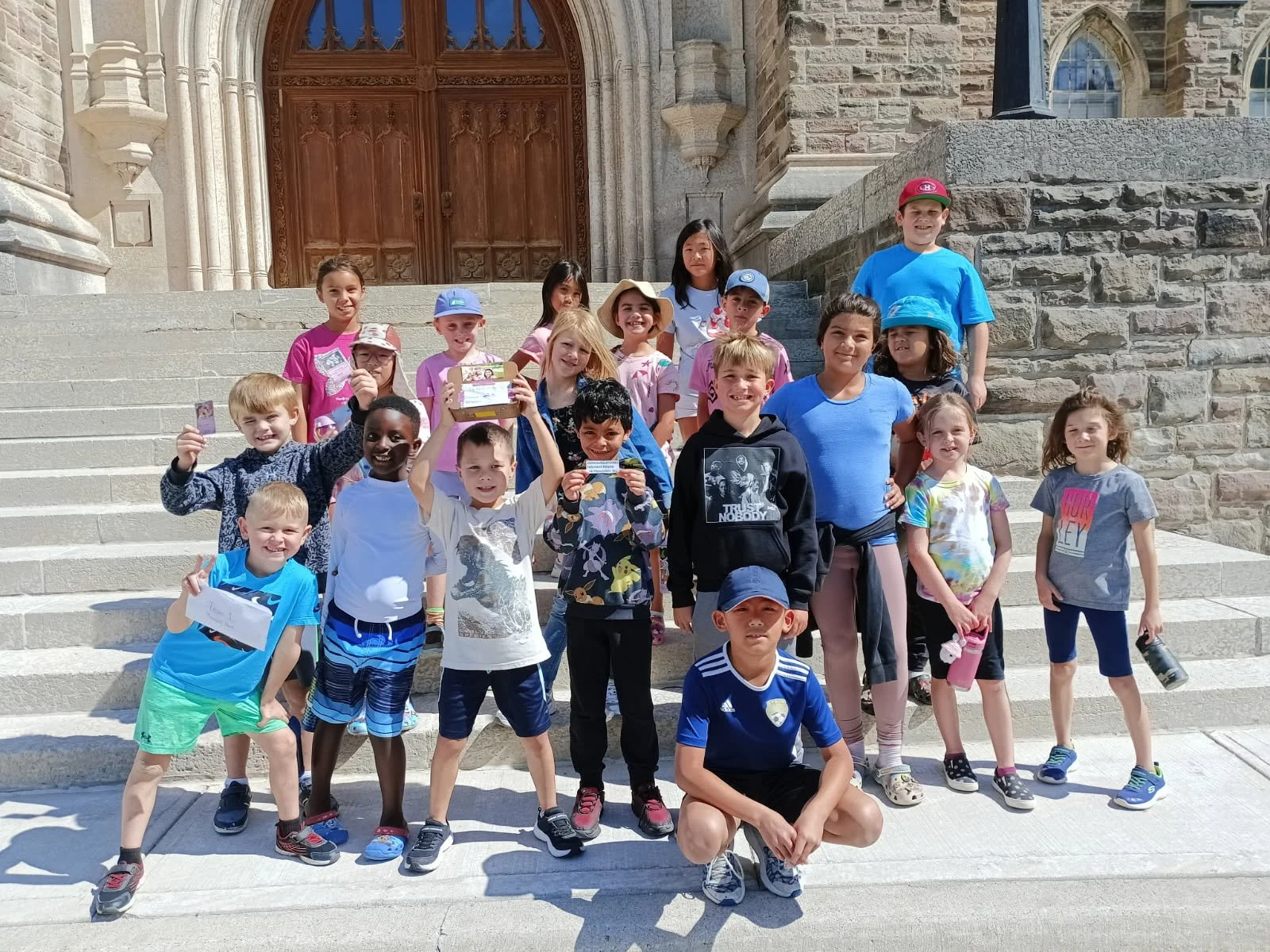Group of young children from Your Neighbourhood Camp standing on steps in front of a building with large wooden door, smiling and holding small items.