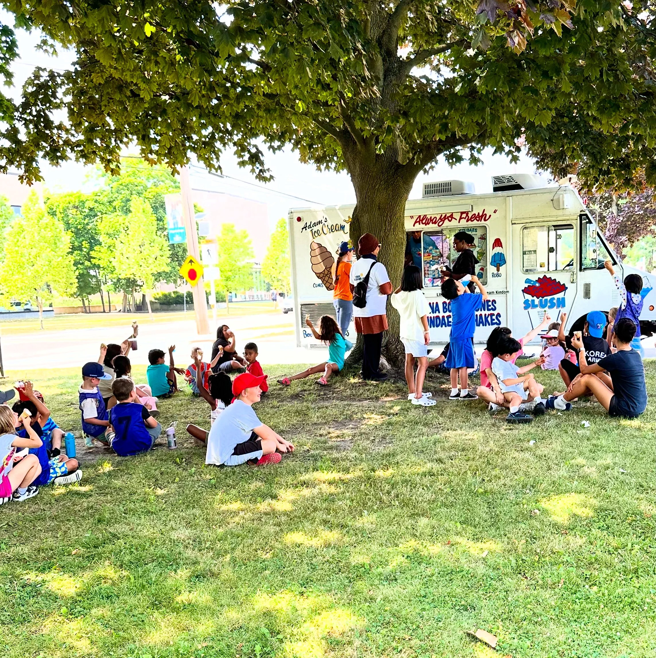 Kids and staff from Your Neighbourhood Camp waiting in line and sitting under a large tree at a colorful ice cream truck in a park on a sunny day.