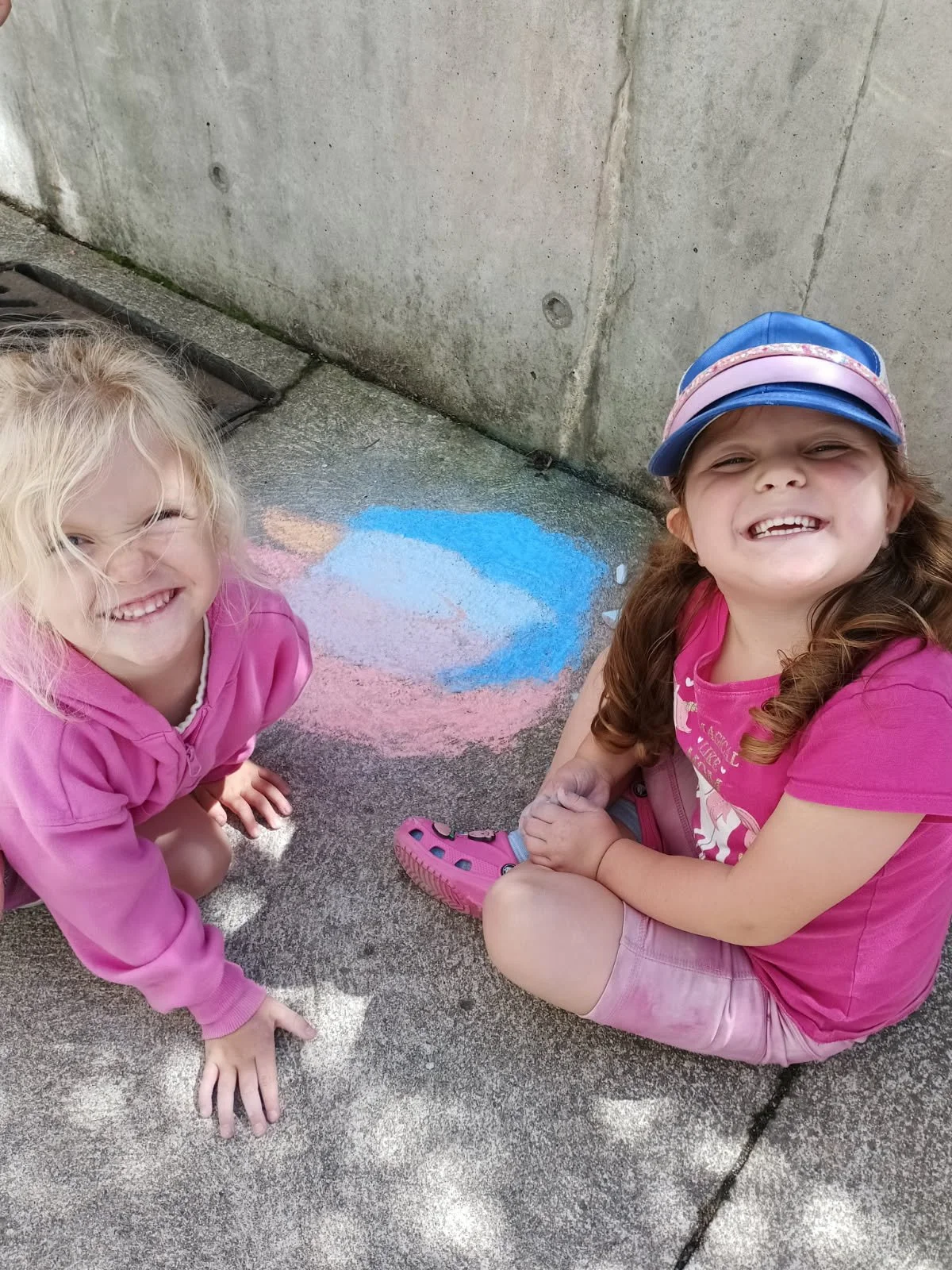 Two young girls at Your Neighbourhood Camp  sitting on a sidewalk near a concrete wall, smiling. There is colorful chalk art of a blue cloud on the ground between them.