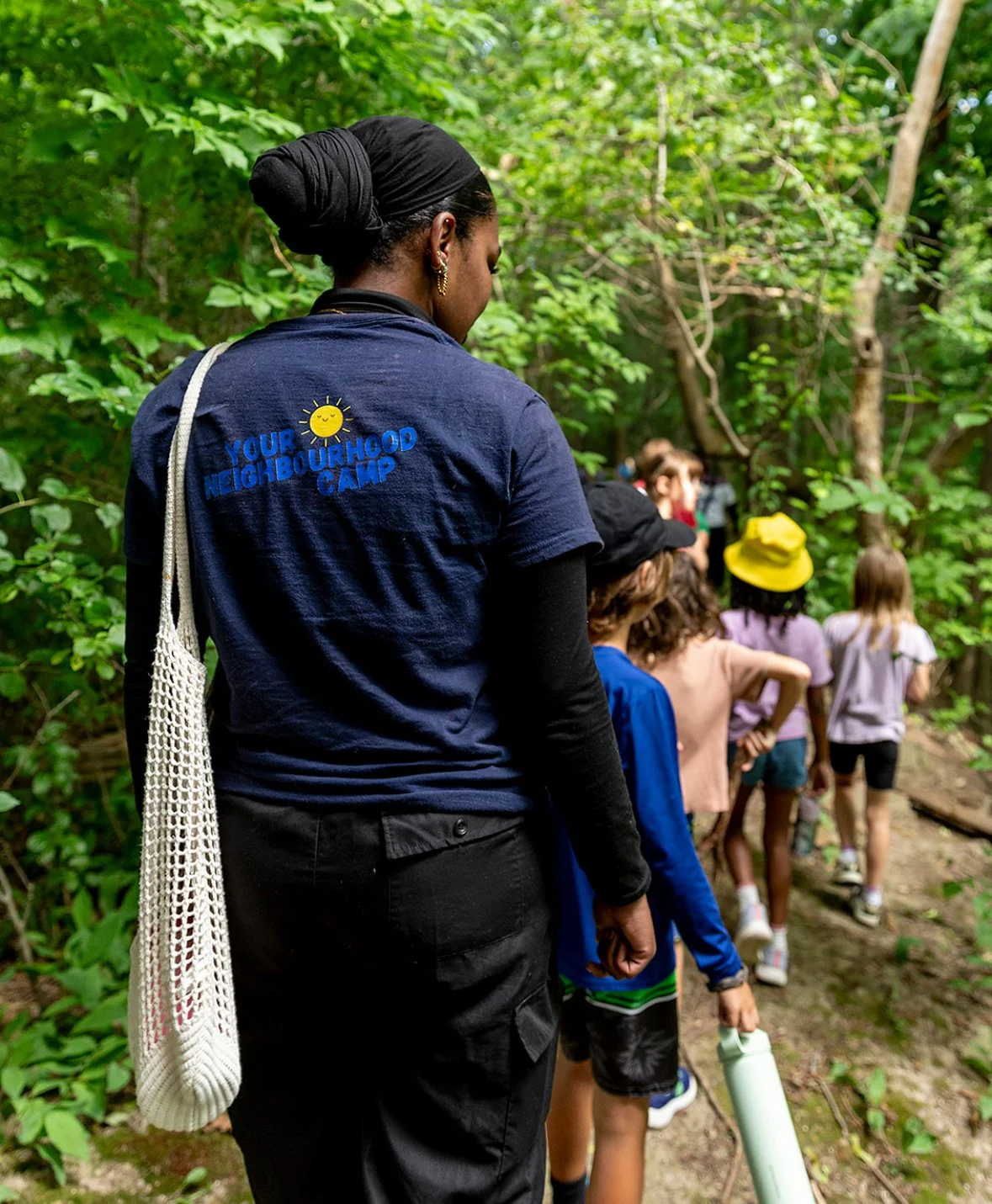 A woman from Your Neighbourhood Camp  with a bundled black hair wrapped in a headscarf, wearing a navy blue T-shirt with a logo and the words 'Your Neighbourhood Camp', stands outdoors among children walking along a wooded trail.