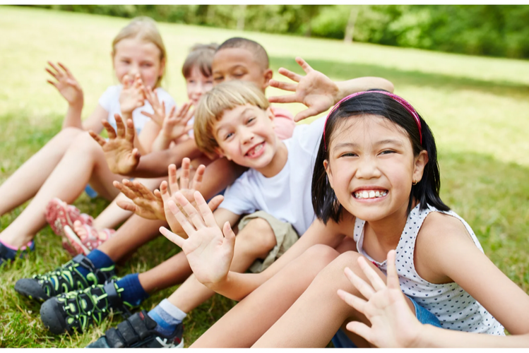 Group of smiling children sitting on grass outdoors, waving and enjoying the moment