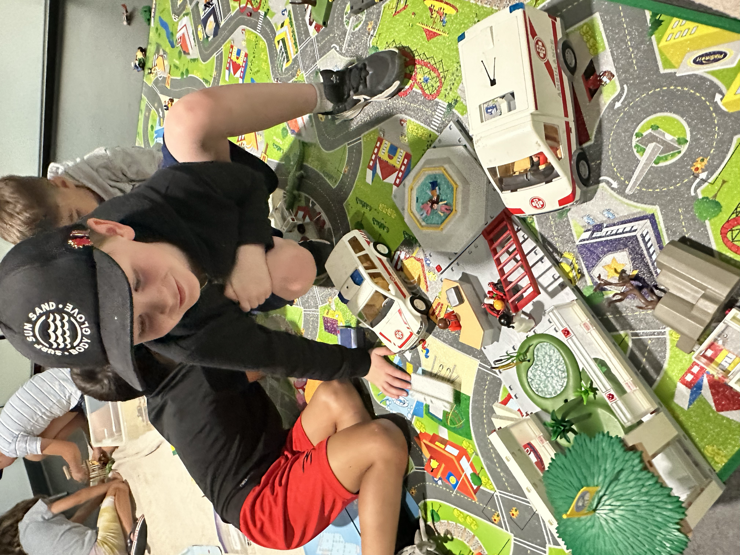 Kids playing at Your Neighbourhood Camp  with a toy city landscape on a colorful mat, with toy vehicles, animals, and buildings.