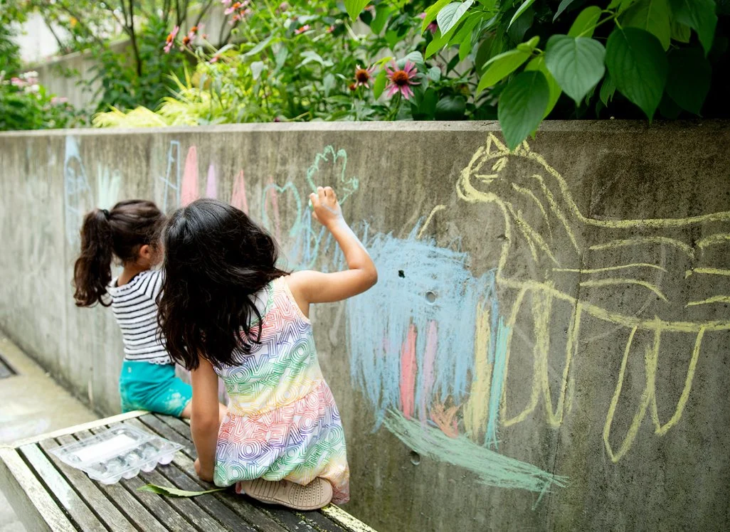 Two young girls at Your Neighbourhood Camp drawing with  colourful chalk art on a concrete wall outdoors, with plants and flowers in the background.