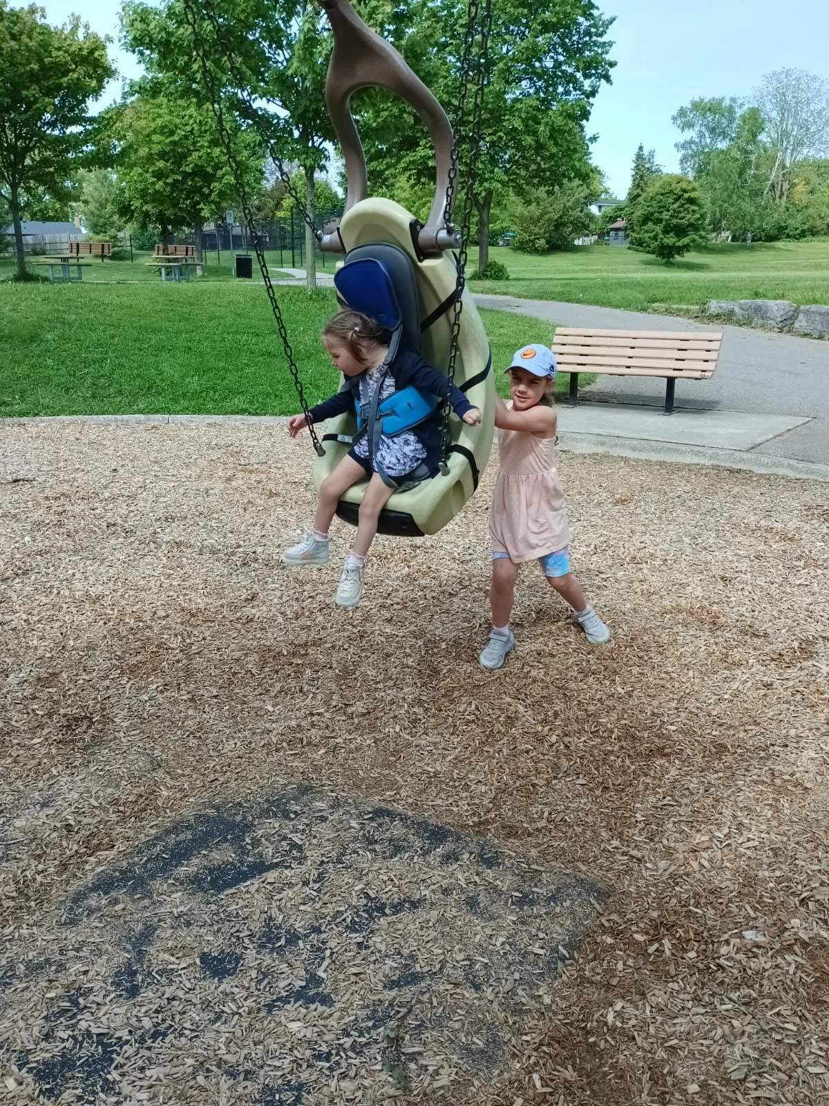 Two young at Your Neighbourhood Camp playing on a swing at a park. One girl is sitting on a dinosaur-shaped swing, and the other girl is pushing it. The park has green trees and grass in the background.