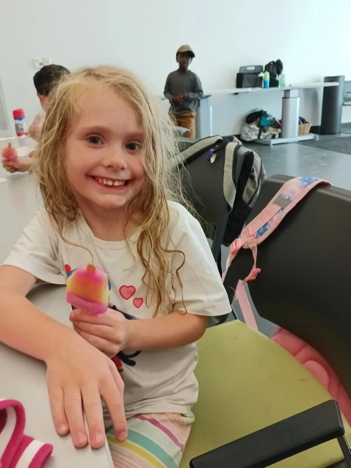 A smiling young girl at Your Neighbourhood Camp with blonde hair, holding a colorful popsicle, sitting at a table in a room with other children and backpacks in the background.
