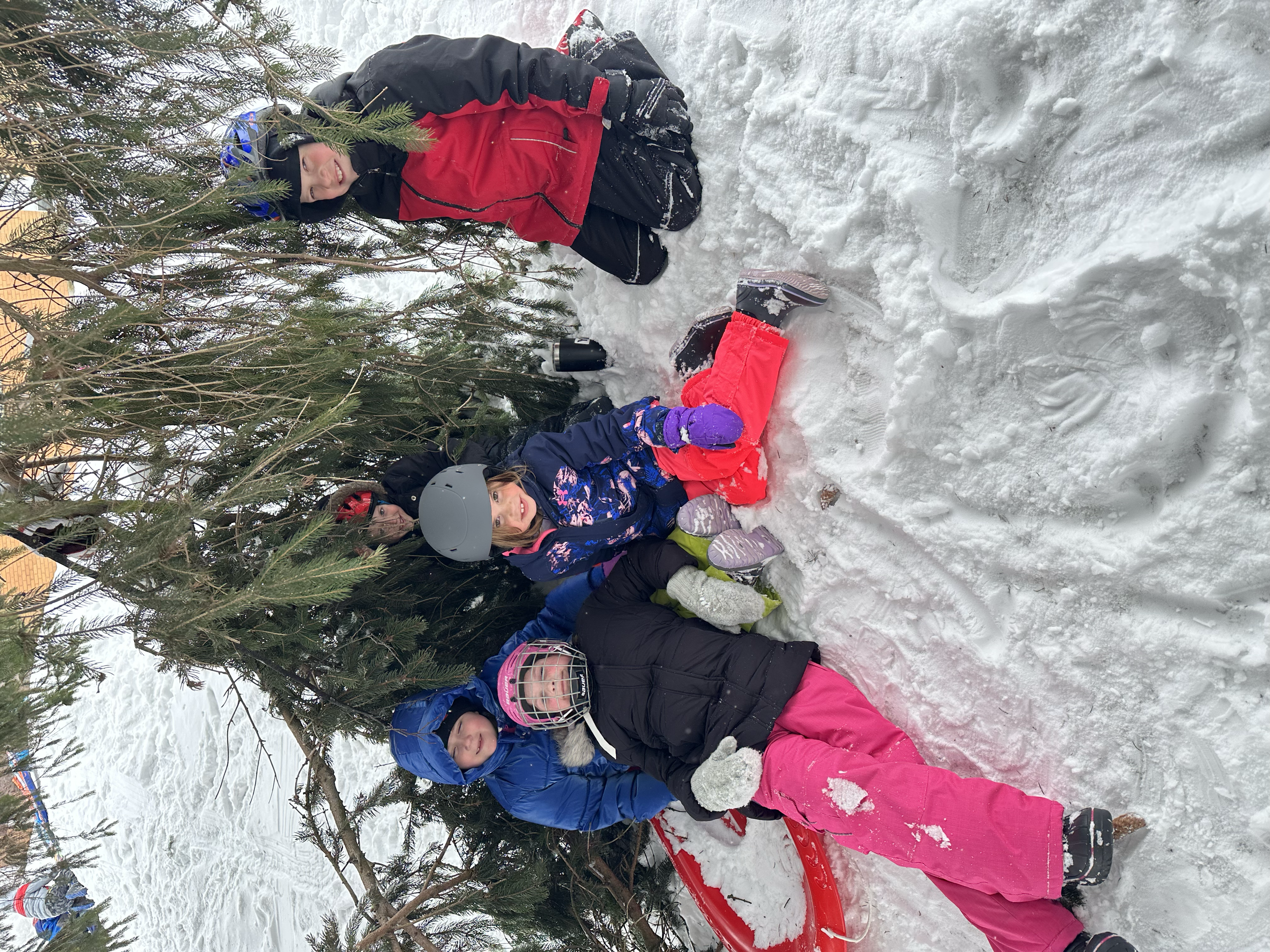 Children and adults at Your Neighbourhood Camp in winter clothing, some on snow sleds, lying and sitting in the snow under a pine tree during daytime, surrounded by snow-covered ground.