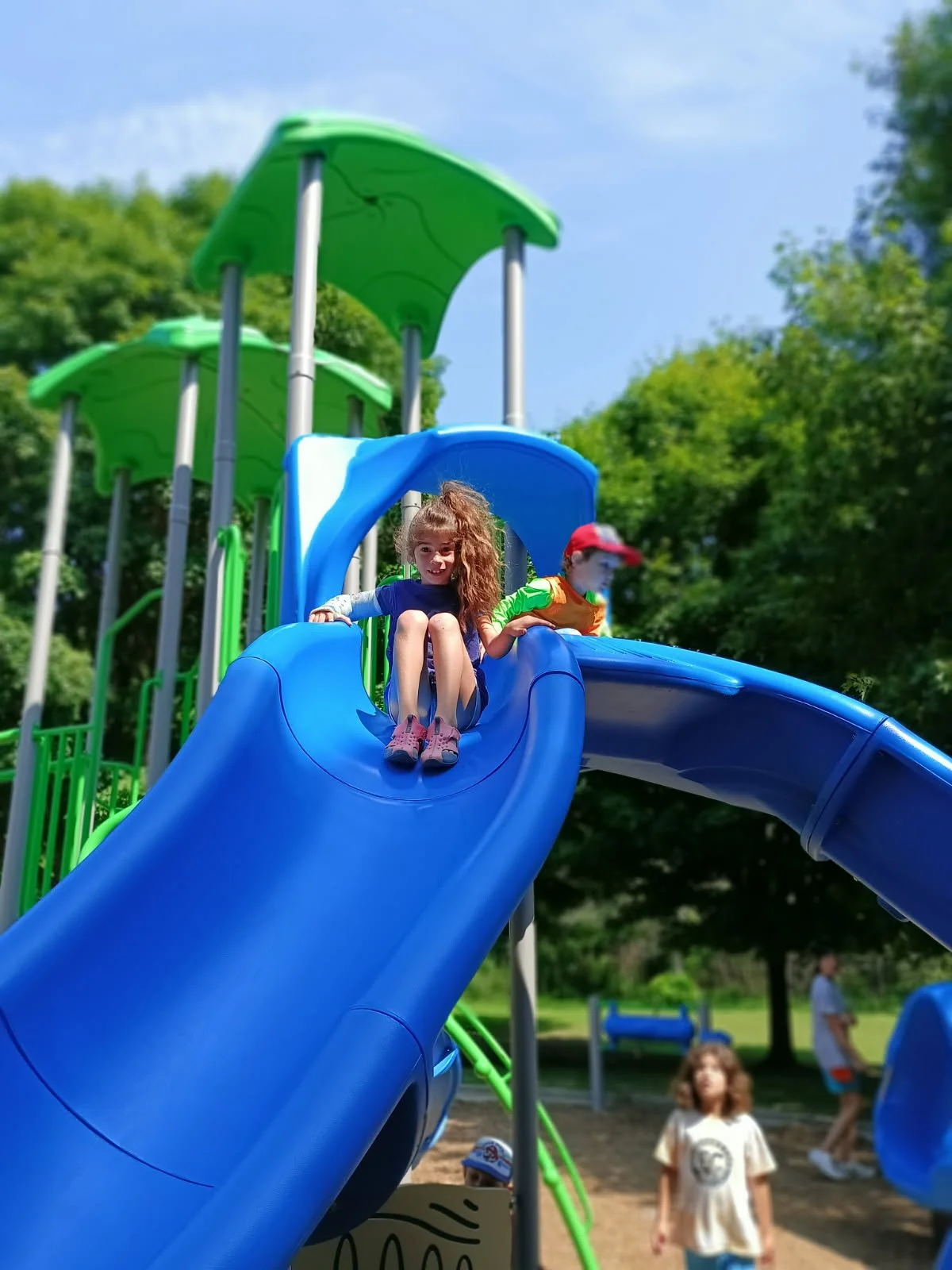 Children  at Your Neighbourhood Camp on a colorful outdoor playground with green and blue slides, surrounded by trees and adult spectators.