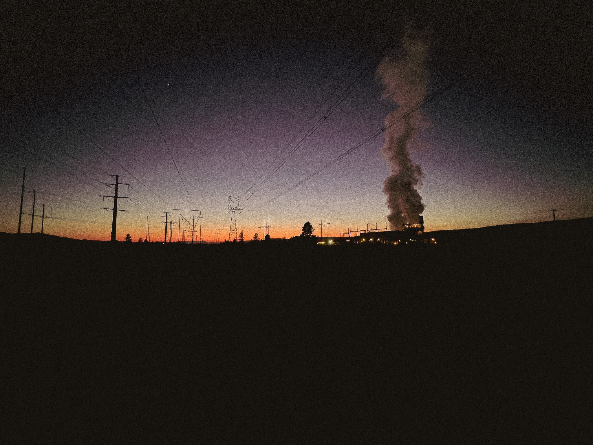 Silhouette of power lines and a factory emitting smoke against a sunset sky