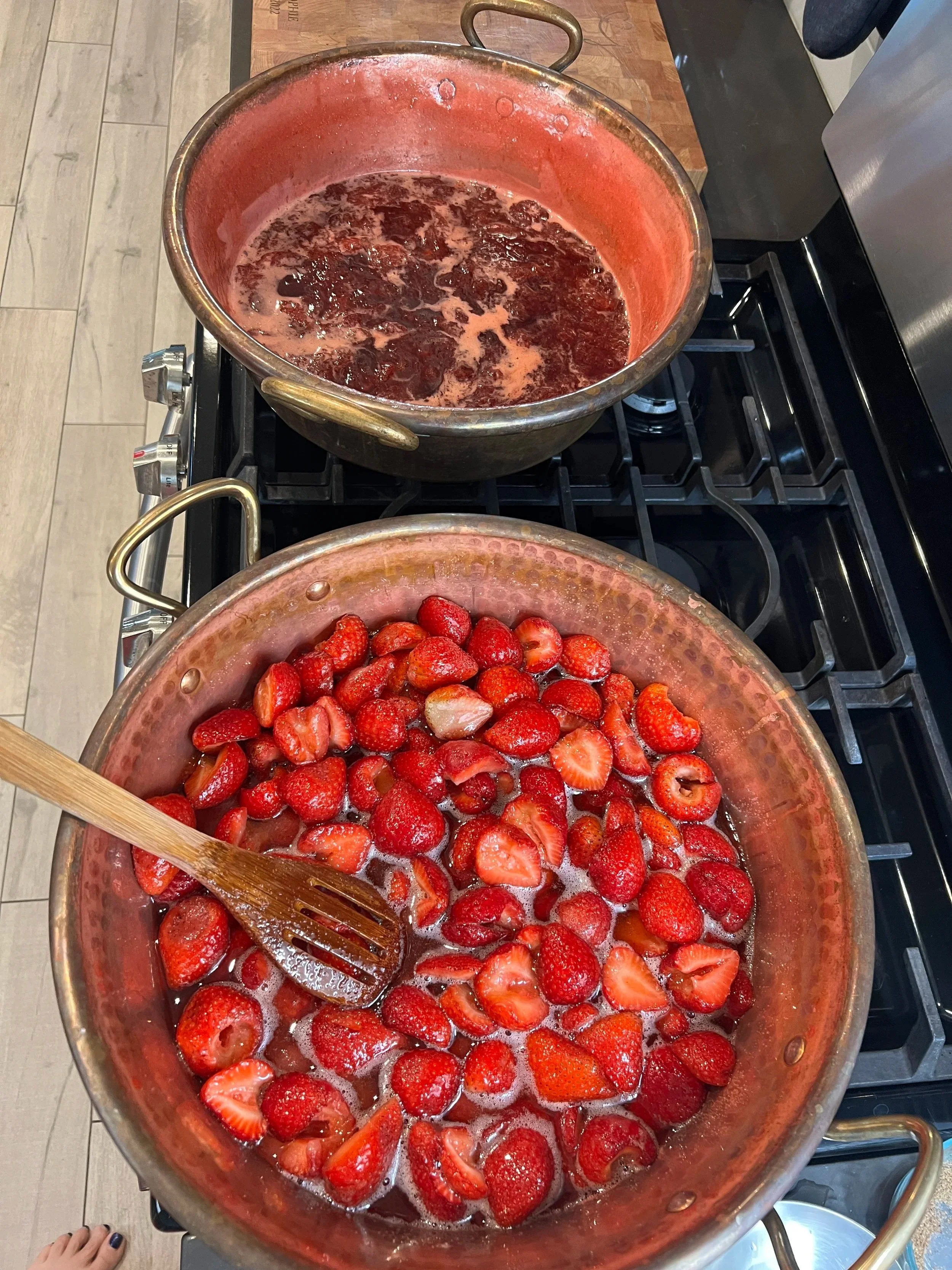 A stainless steel pot on a stove filled with sliced strawberries cooking, with bubbles forming on the surface. A wooden spoon rests in the pot.