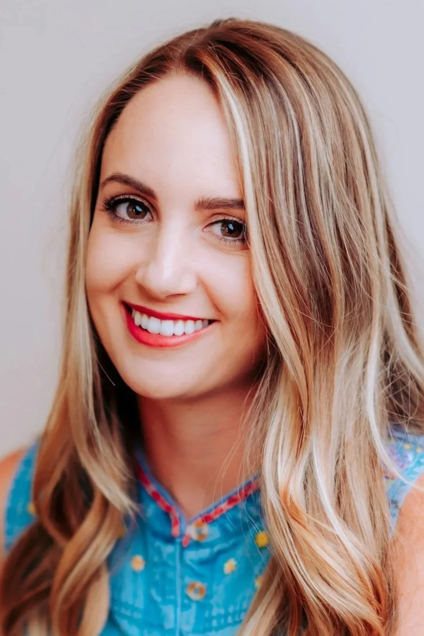 Close-up of a young woman with long, wavy blonde hair, smiling, wearing a blue sleeveless top with floral embroidery.