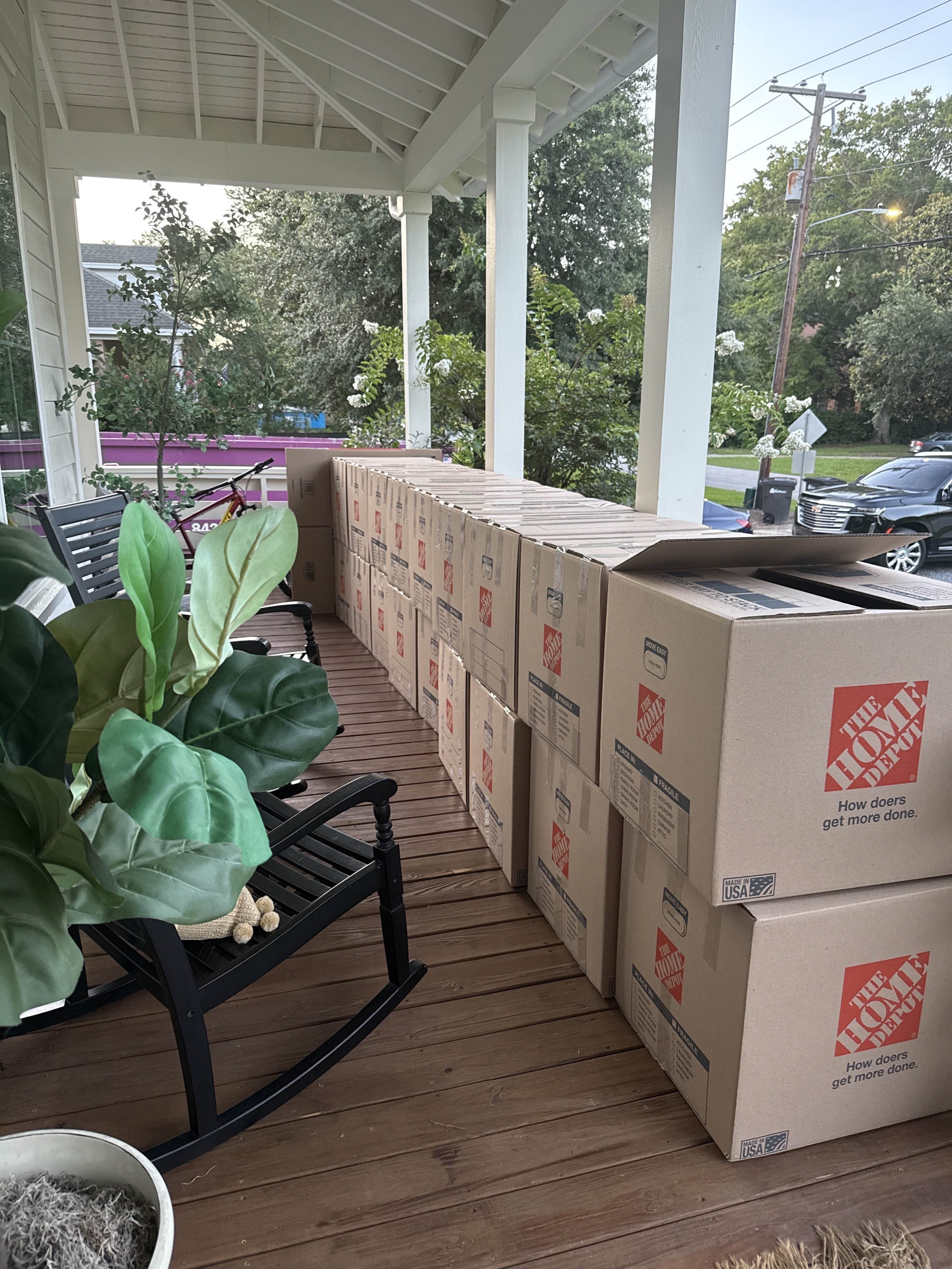 Multiple cardboard boxes of The Home Depot, stacked outside on a porch with plants and chairs.