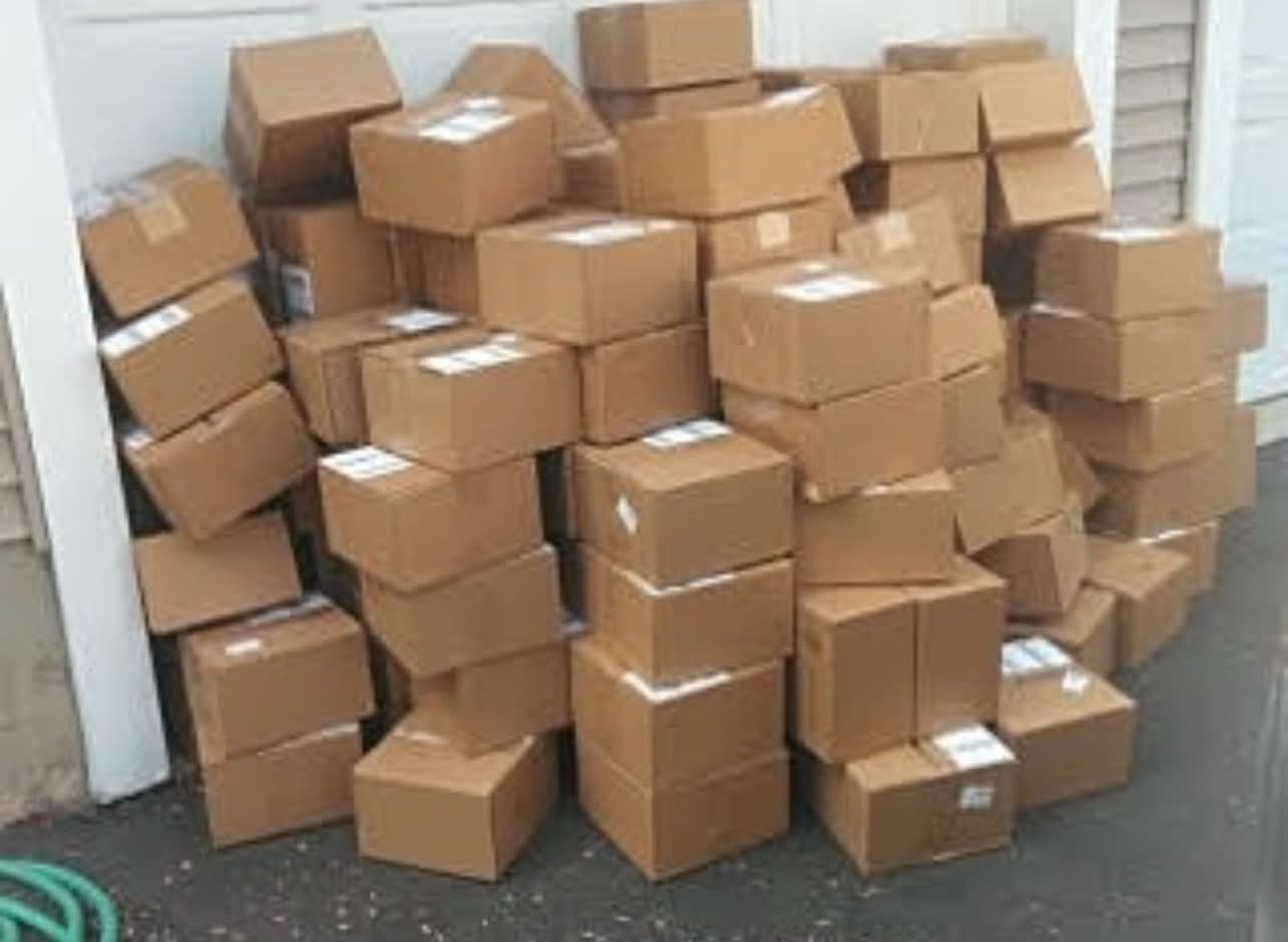 Multiple cardboard boxes stacked outside on a gray surface, some leaning against a building wall.