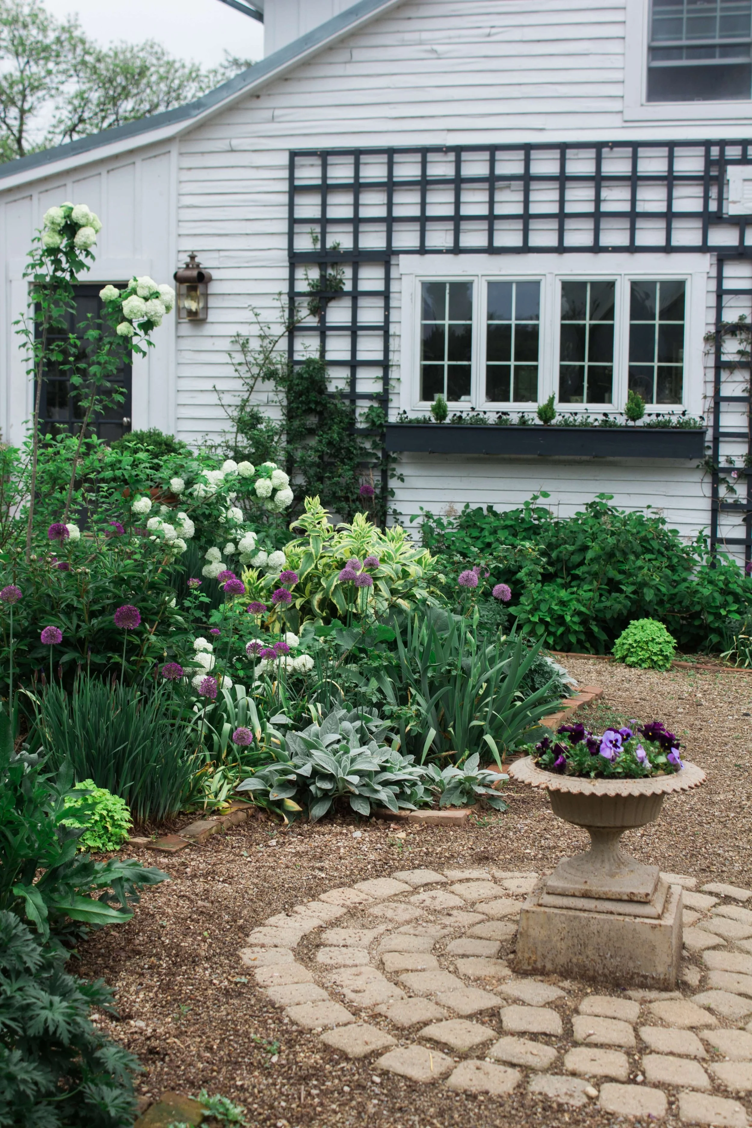 Jardín con flores y plantas frente a una casa blanca con ventanas, en una parcela de tierra con paso de piedras, y un florero con flores en primer plano.