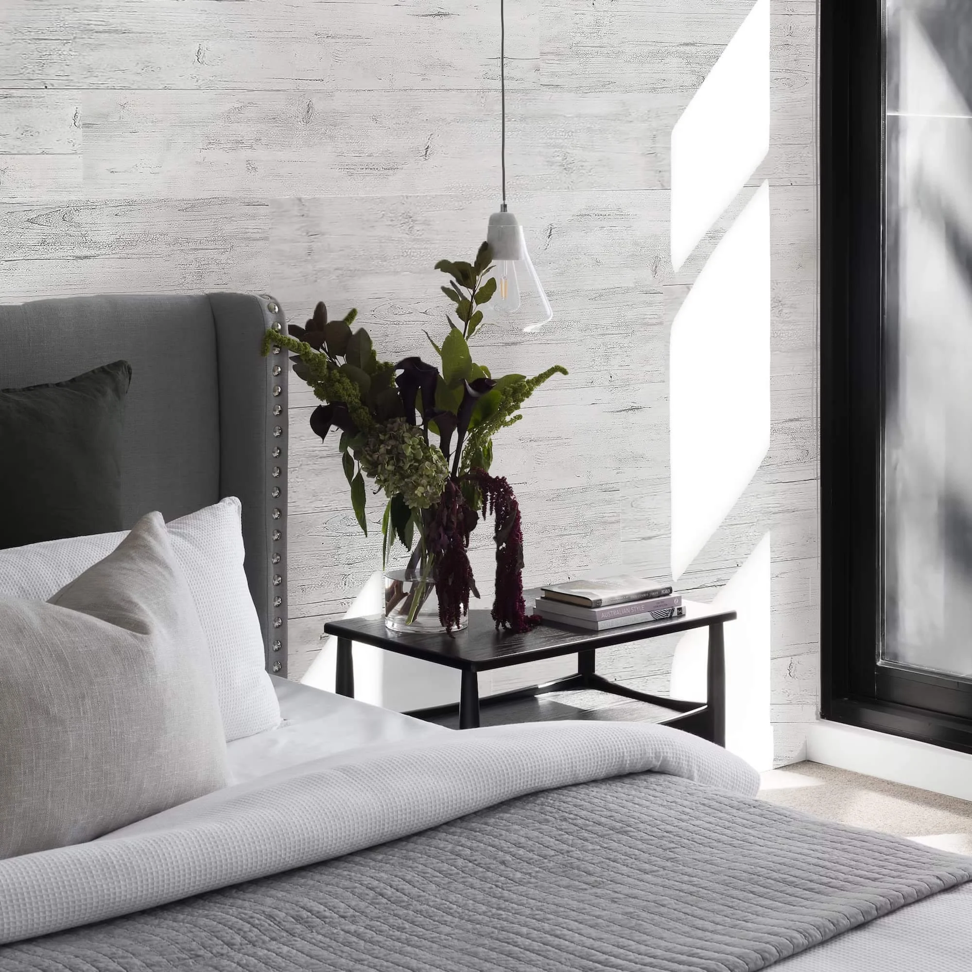 Habitación con cama con sábanas blancas y cojines beige y gris. Refrigerador negro en la esquina. Ventana grande con luz natural, pared de madera pintada de blanco. Mesa con jarrón con flores y libros al lado.