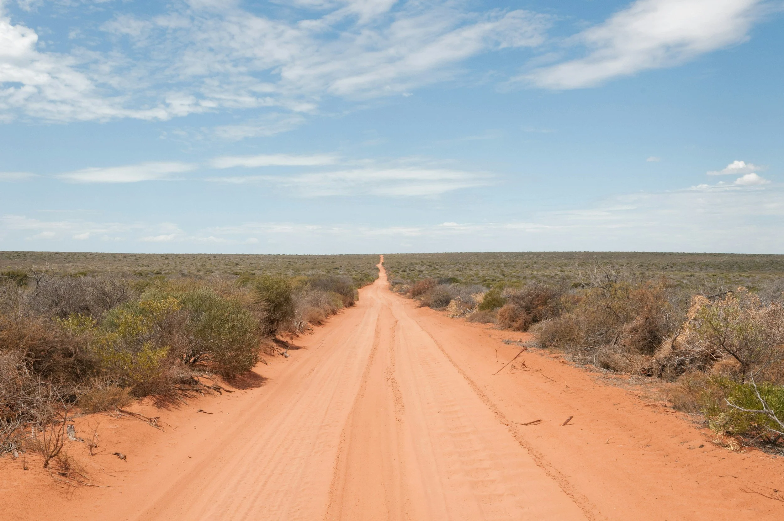 A dirt road stretching straight through a flat, arid landscape with sparse desert vegetation, under a partly cloudy blue sky.