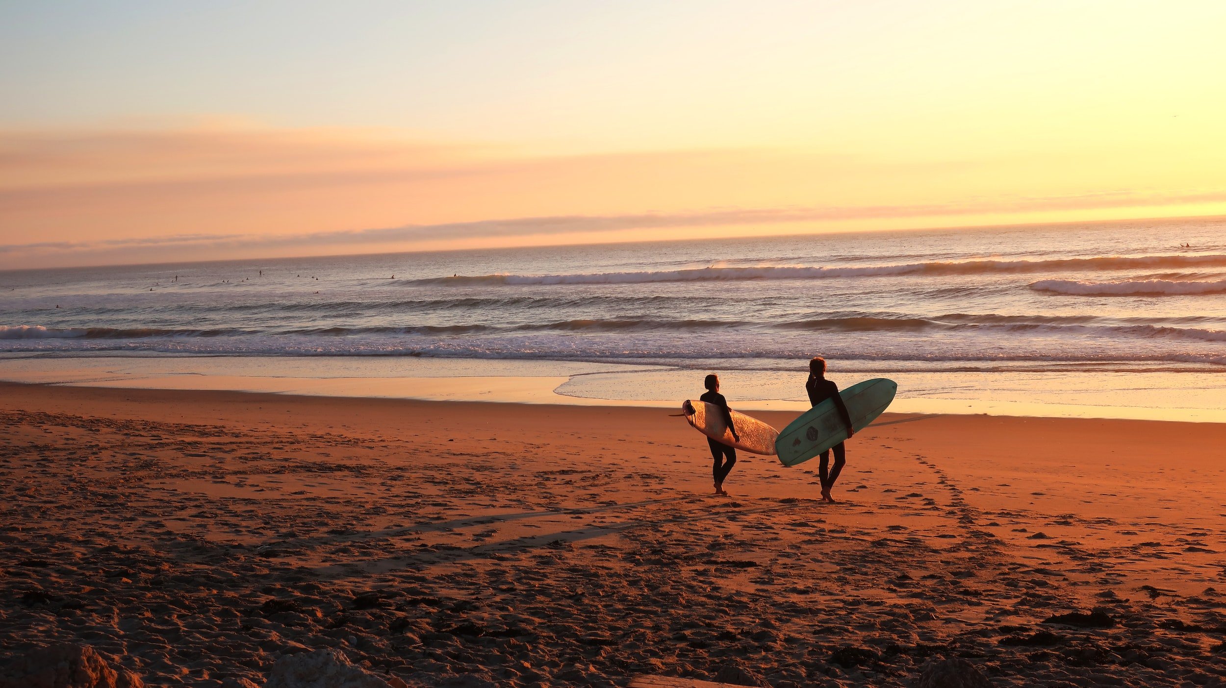 Two surfers walking on the beach at sunset, carrying surfboards with the ocean waves in the background.