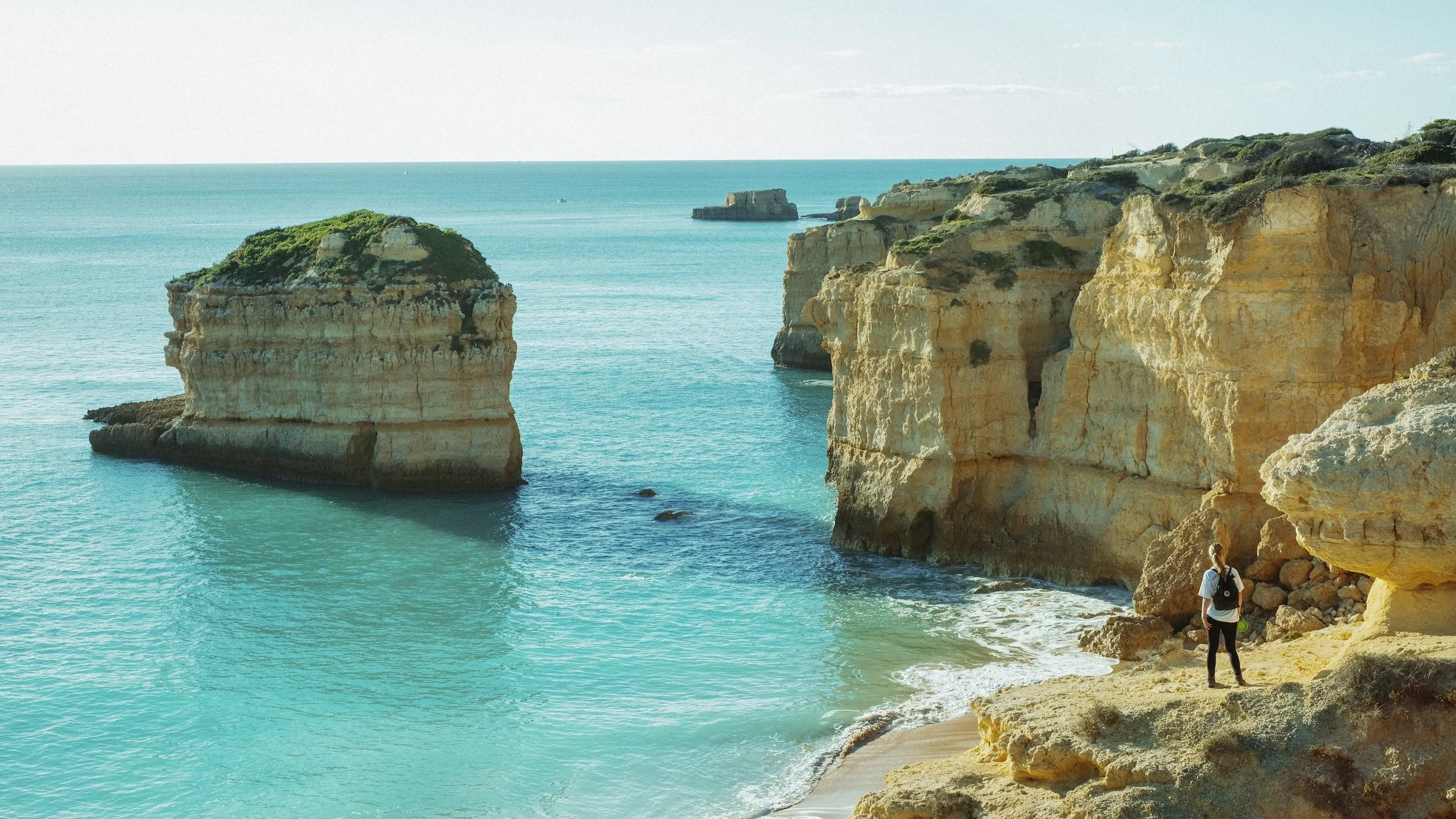 Cliffs and rock formations along a blue ocean, with a person standing on the rocky shore wearing a backpack and looking at the scenery.