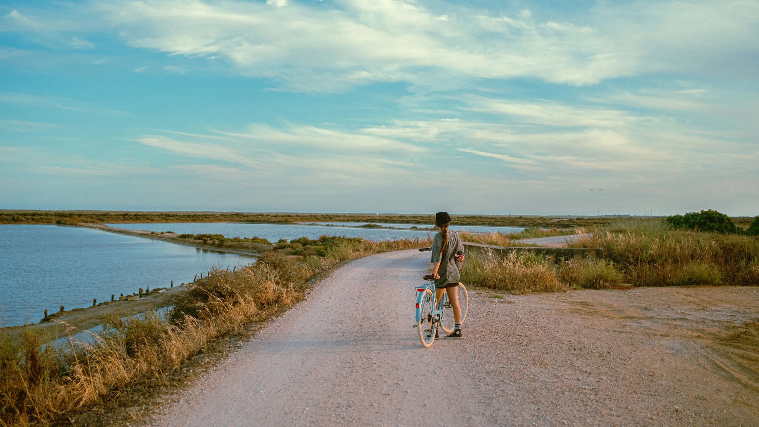 Person riding a bicycle on a gravel path by a body of water under a partly cloudy sky.