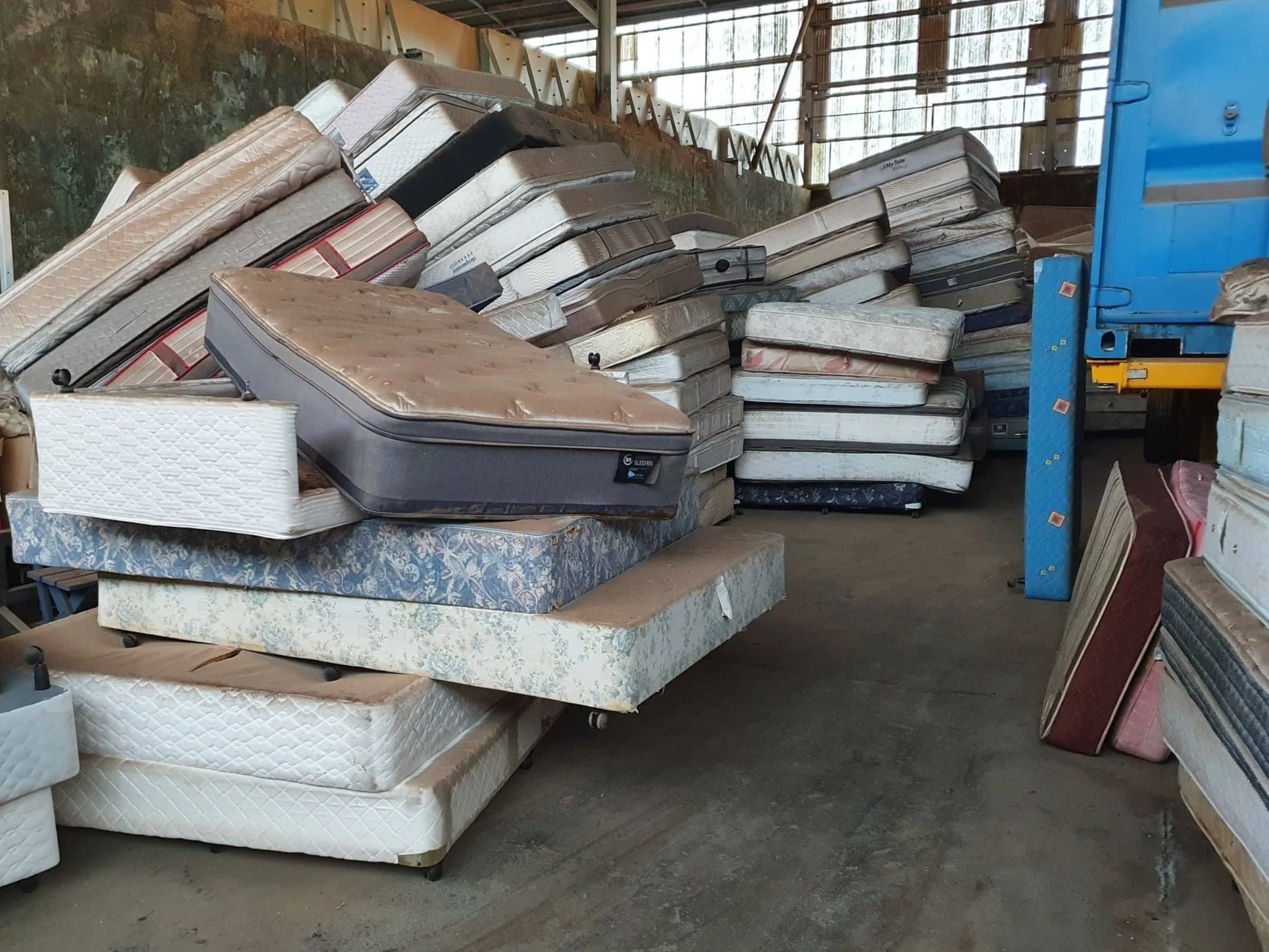 A large pile of old, used mattresses stacked haphazardly on the floor inside a warehouse awaits mattress recycling.