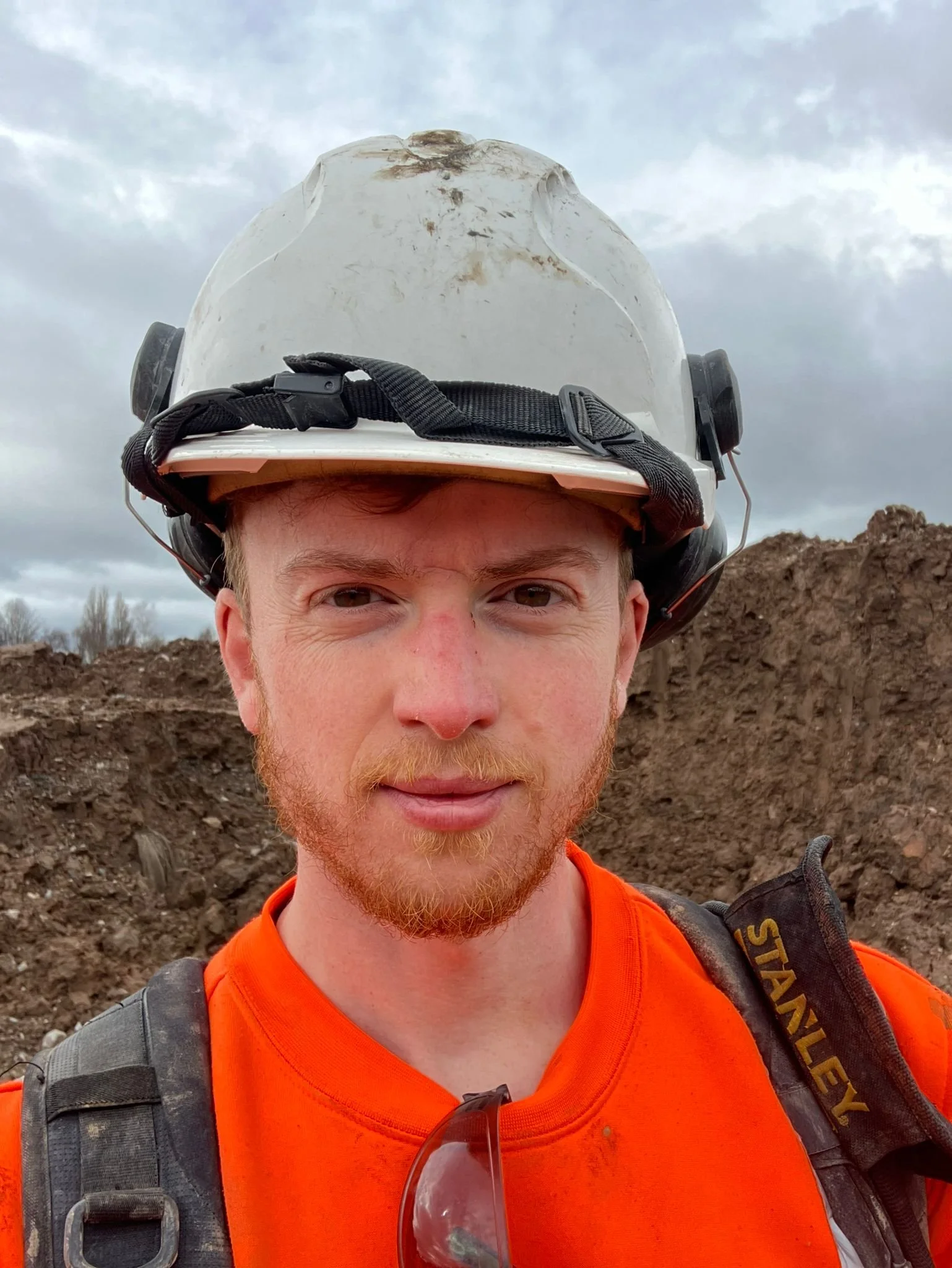 Construction worker with a white safety helmet and orange shirt standing outdoors with dirt mounds and cloudy sky in background.