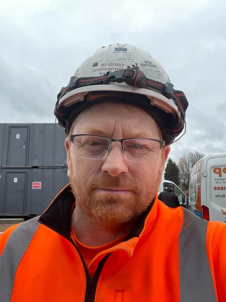 A construction worker wearing a safety helmet, glasses, and an orange safety vest, standing outdoors with a cloudy sky, industrial equipment, and vehicles in the background.