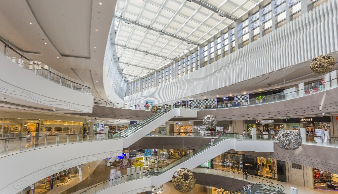 Interior view of a modern shopping mall with multiple levels, escalators, and a glass ceiling letting in natural light.