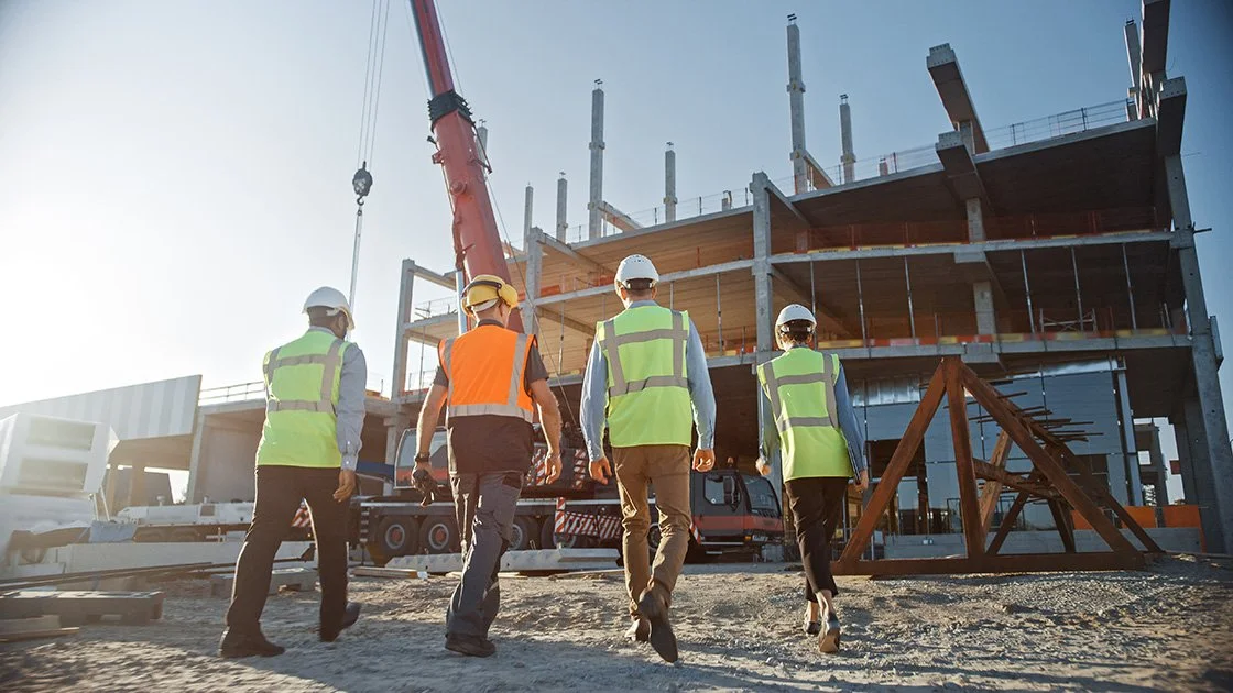Construction site with five workers wearing safety vests and helmets, overseeing a building under construction with a crane in the background.
