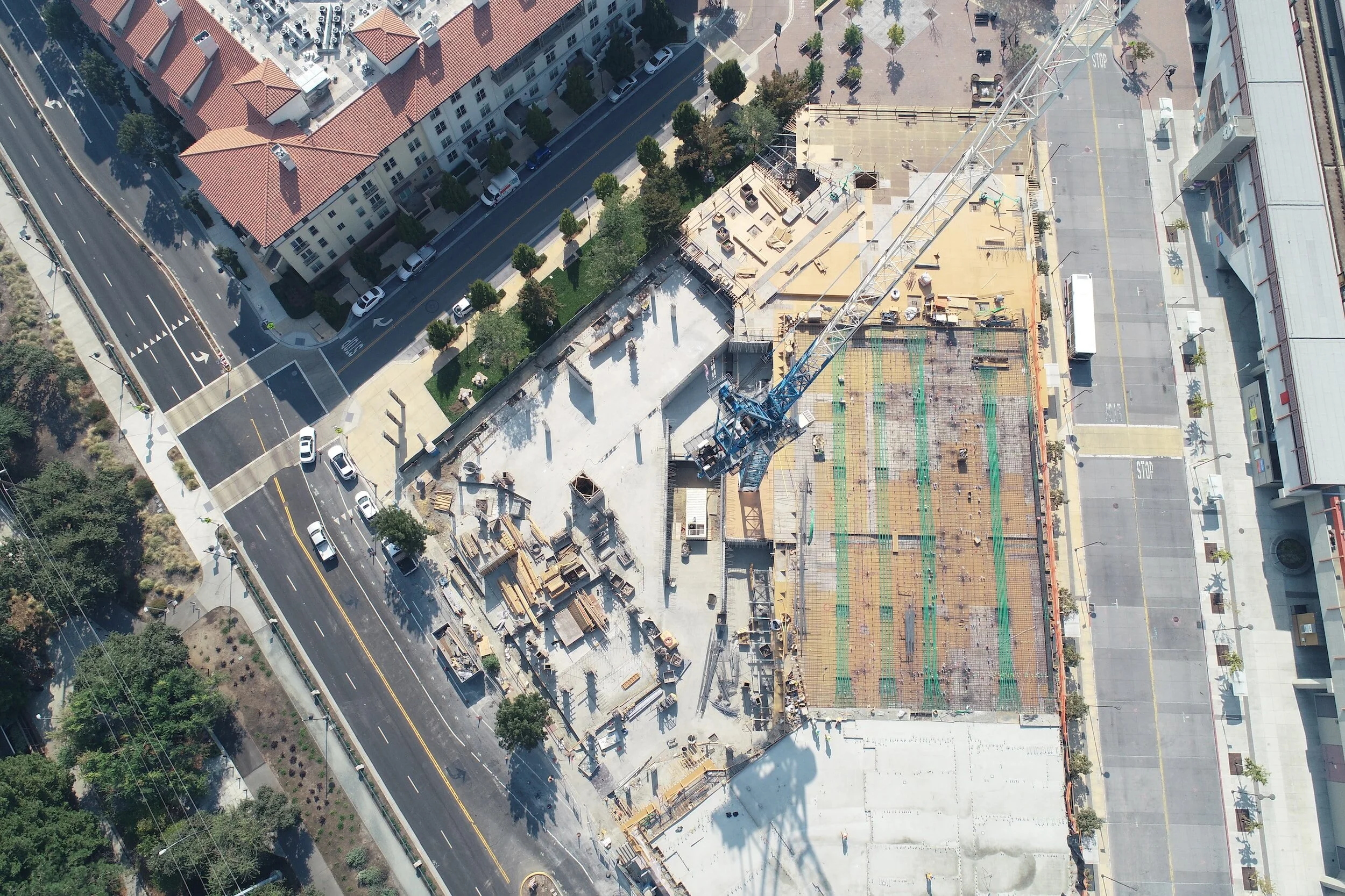 Aerial view of a construction site with a crane, surrounded by residential and commercial buildings, streets, and parking lot