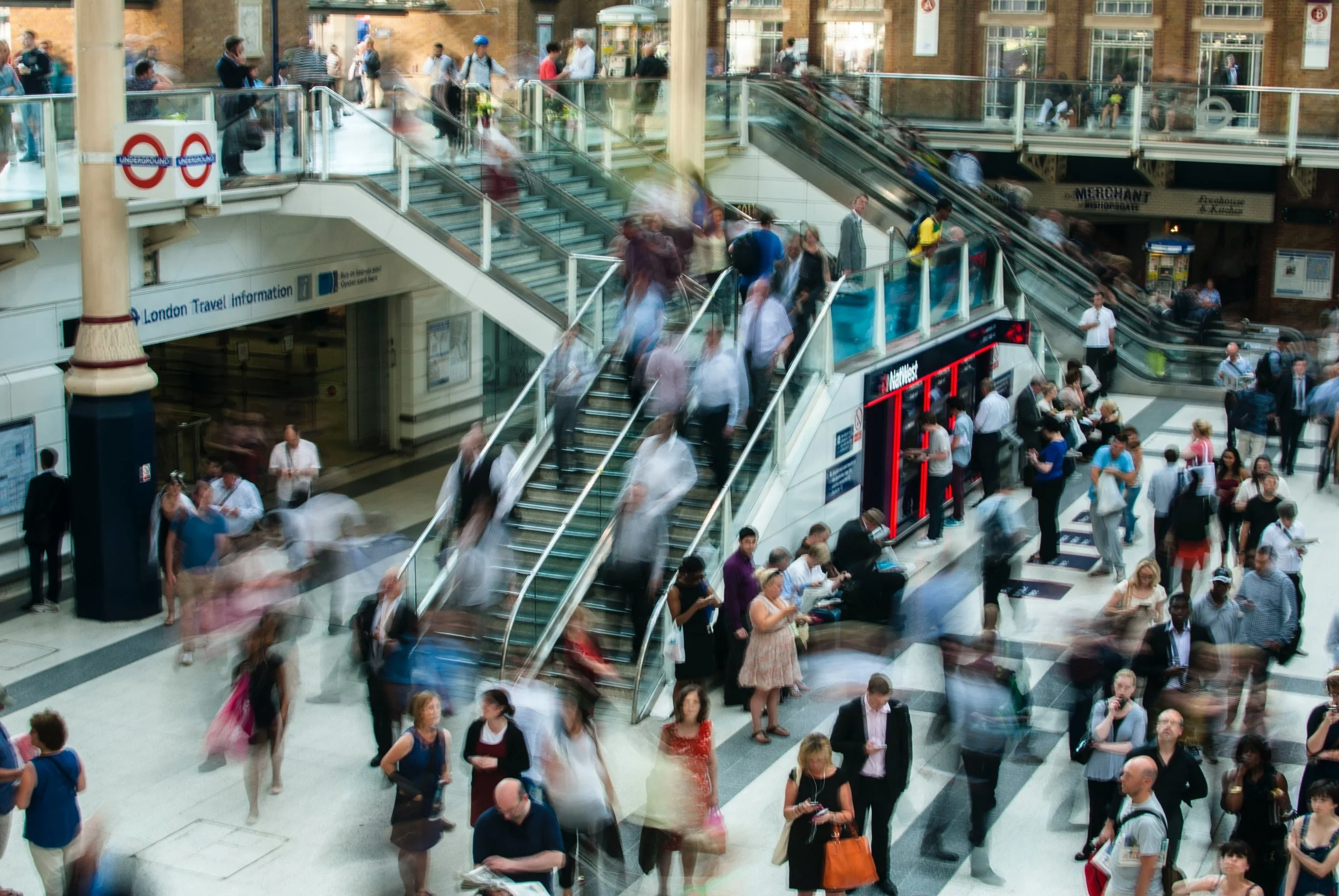 Crowded underground train station with people ascending and descending stairs, walking, talking, and waiting.