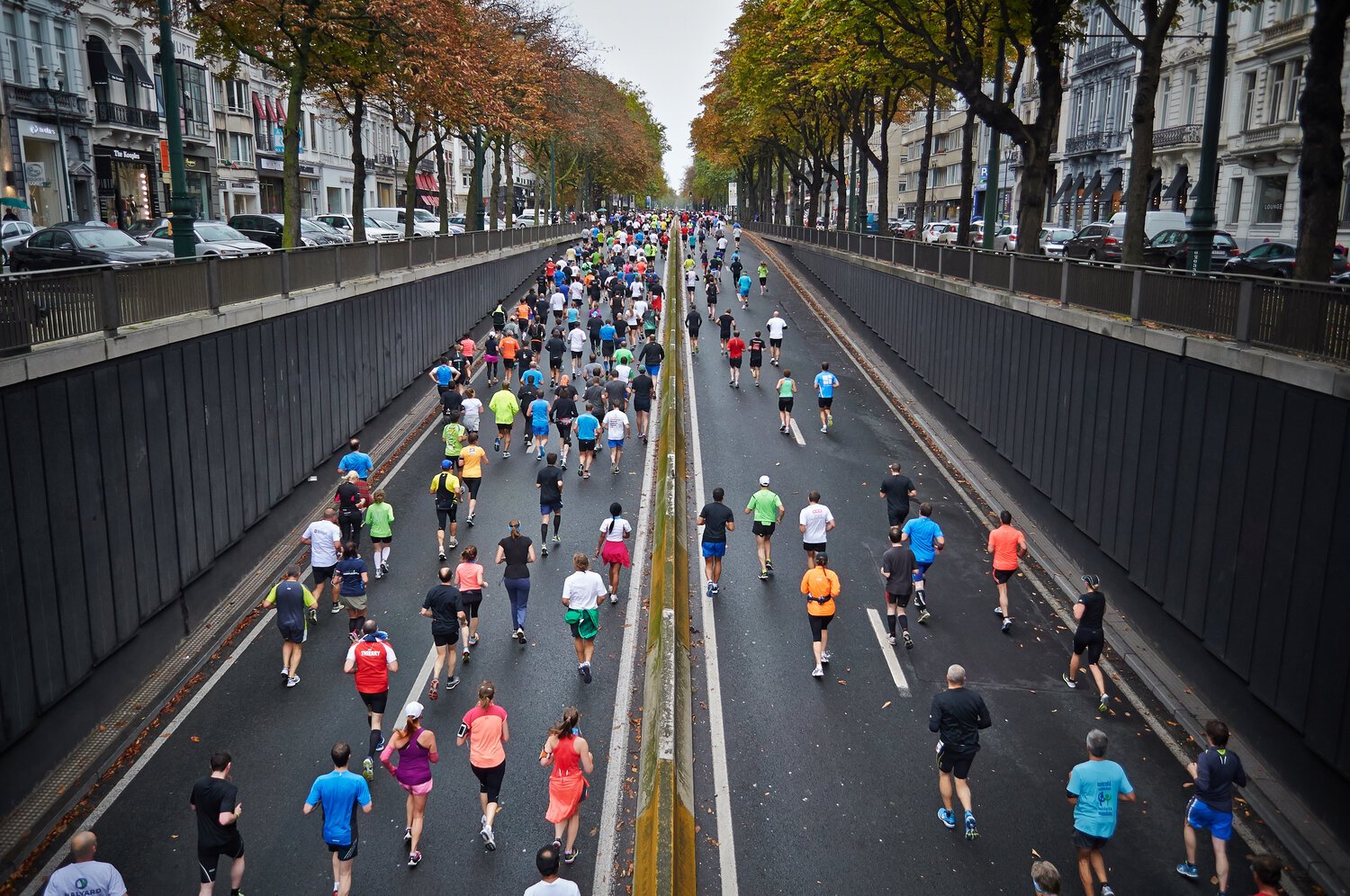 Crowd of people participating in a running event or marathon on a city street, with trees and buildings lining both sides.