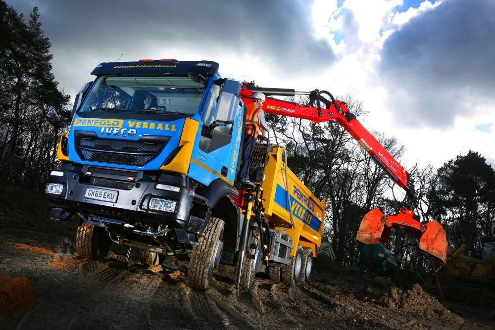 A construction truck with a long red lifting arm and orange bucket, working on a dirt site with trees and cloudy sky in the background.