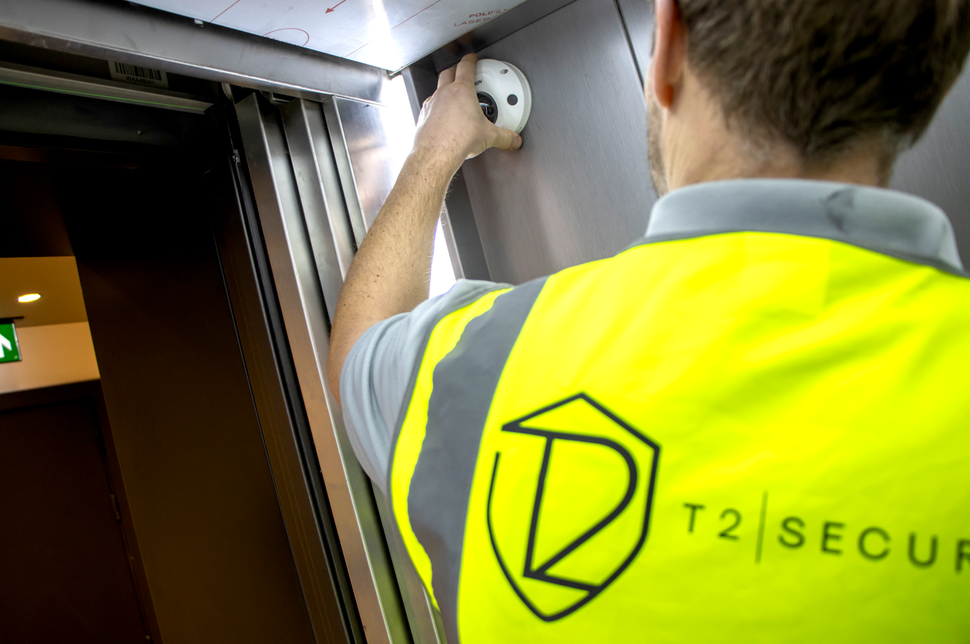 A security personnel in a yellow reflective vest with a shield logo and the words "T2 Secure" is working with a security camera in an airport or train station.