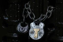 Close-up of a black chain and padlock with a silver heart-shaped lock that has a gold letter 'T' on it.