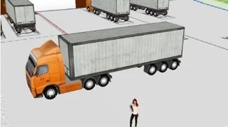A person standing near an orange semi-truck with a grey trailer in a parking lot with multiple trucks in the background.