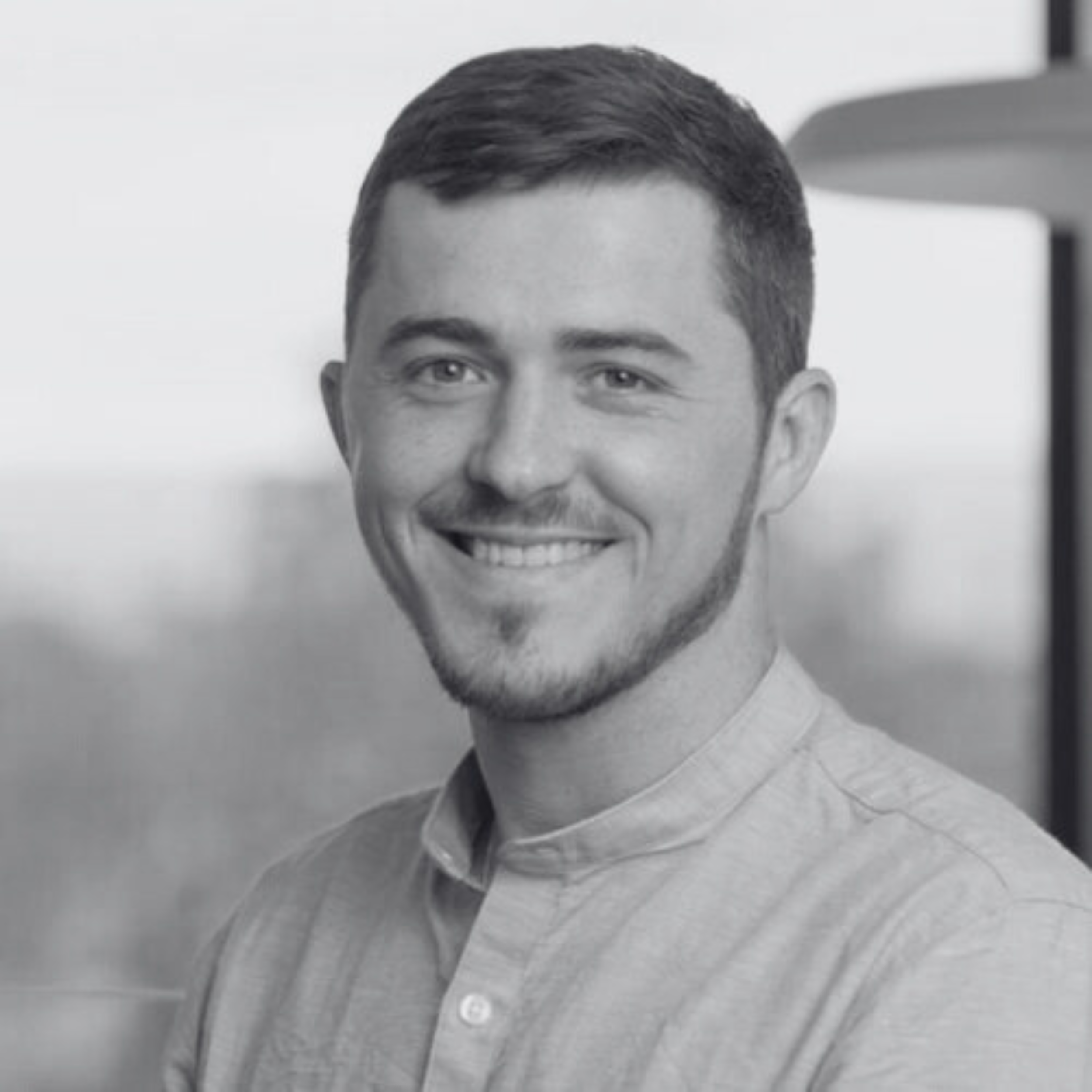 Black and white portrait of a young man with short dark hair, light skin, and a slight beard, smiling and looking at the camera, wearing a collared shirt.