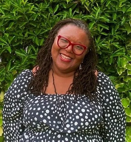 Woman with curly hair wearing red glasses and a black and white polka dot dress, standing in front of green bushes.