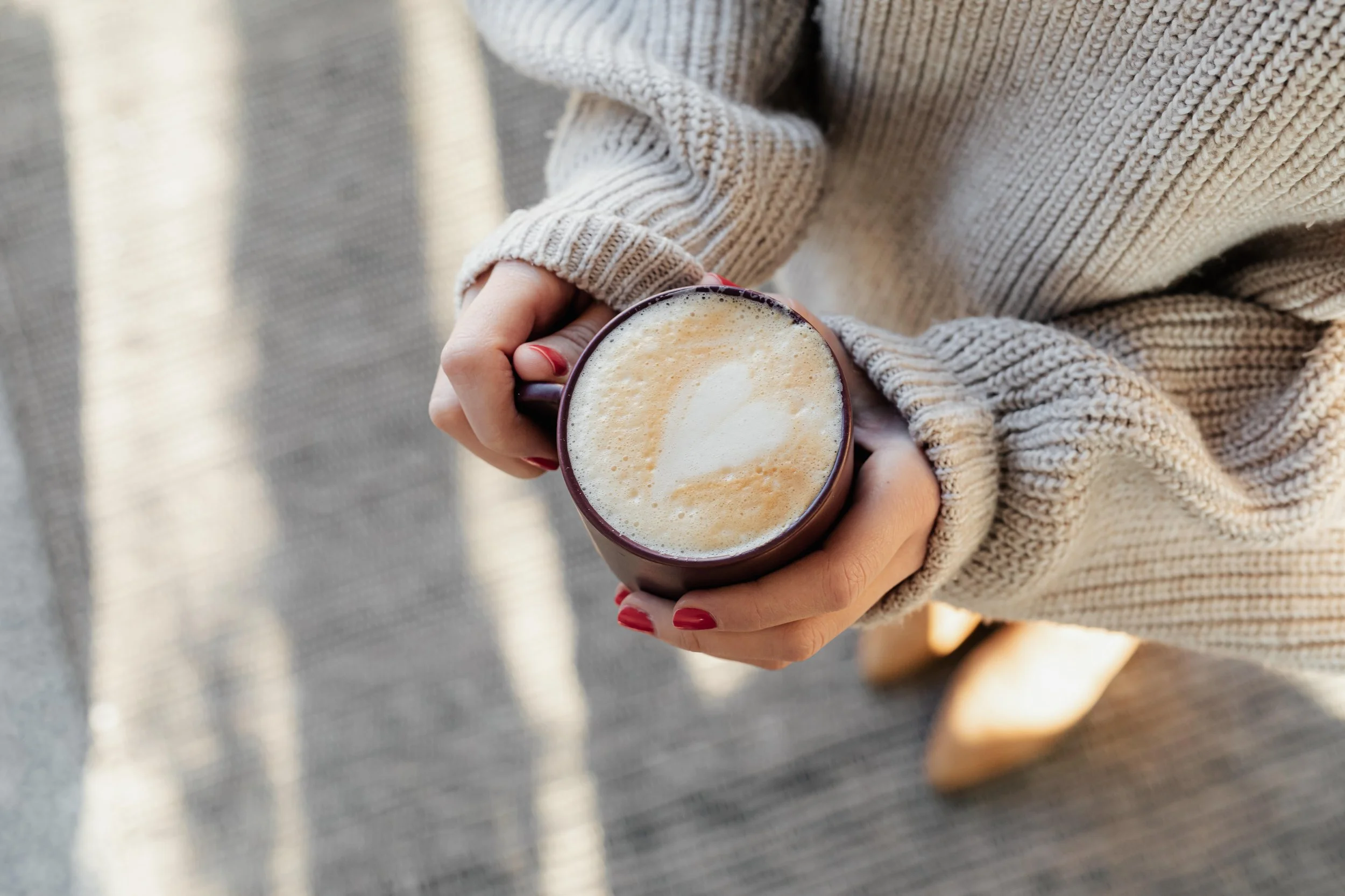 Person hält eine Tasse Cappuccino mit Milchschaum und Herzdekoration. Die Person trägt einen beige Strickpullover und hat rote Nagellacke. Im Hintergrund ist ein grauer Teppich sichtbar.