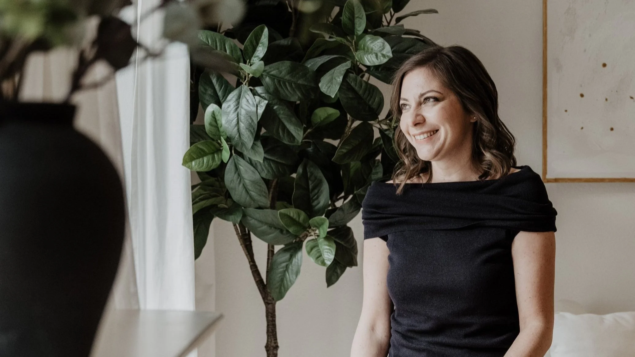 Eine lachende Frau mit braunen, mittellangen, gewellten Haaren in einem schwarzen Kleid sitzt neben einem großen grünen Zimmerpflanzen in einem hellen Raum.